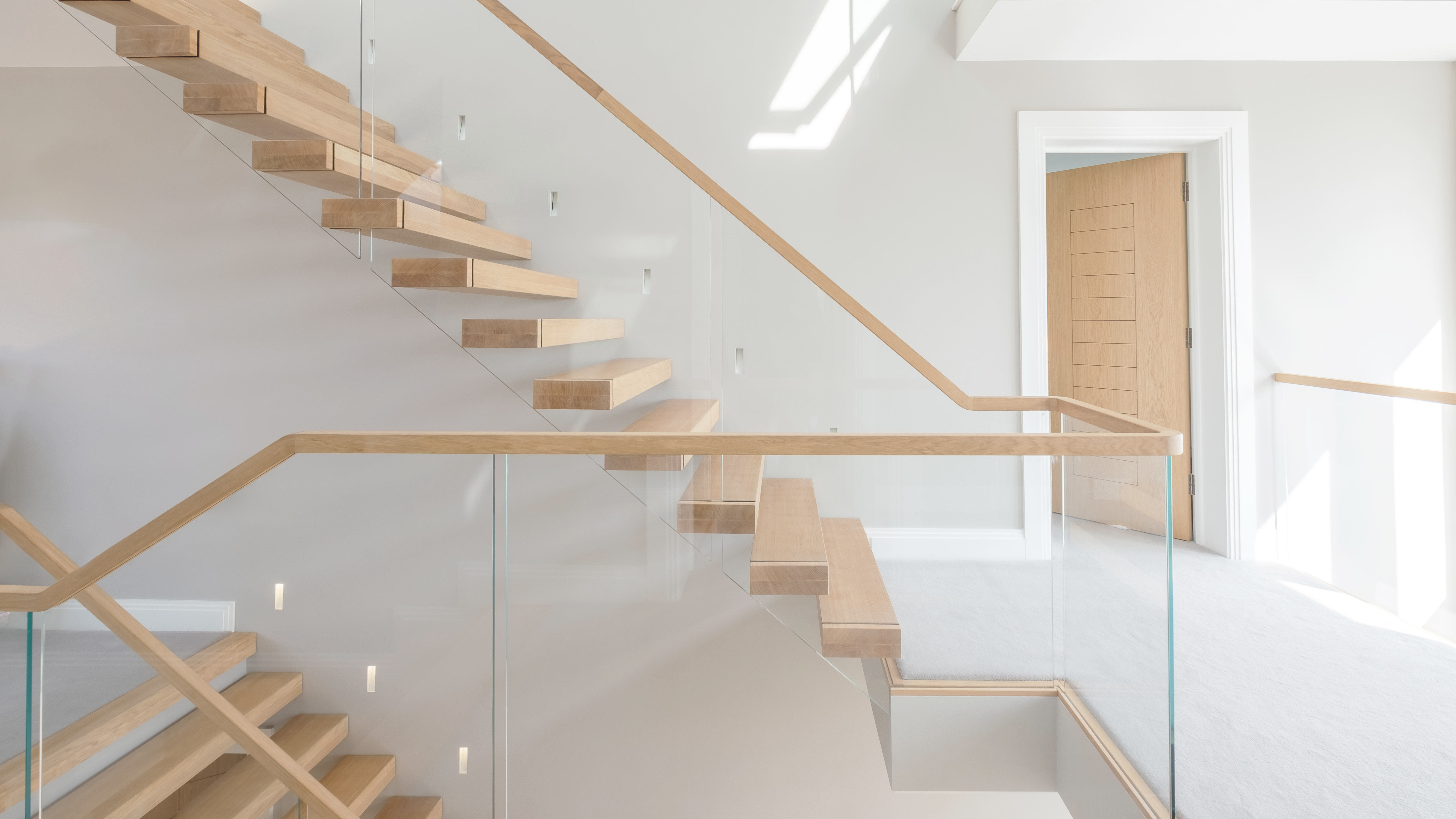Side view of cantilevered floating oak stair over three stories with glass balustrade. Grey walls and carpet with oak doors.