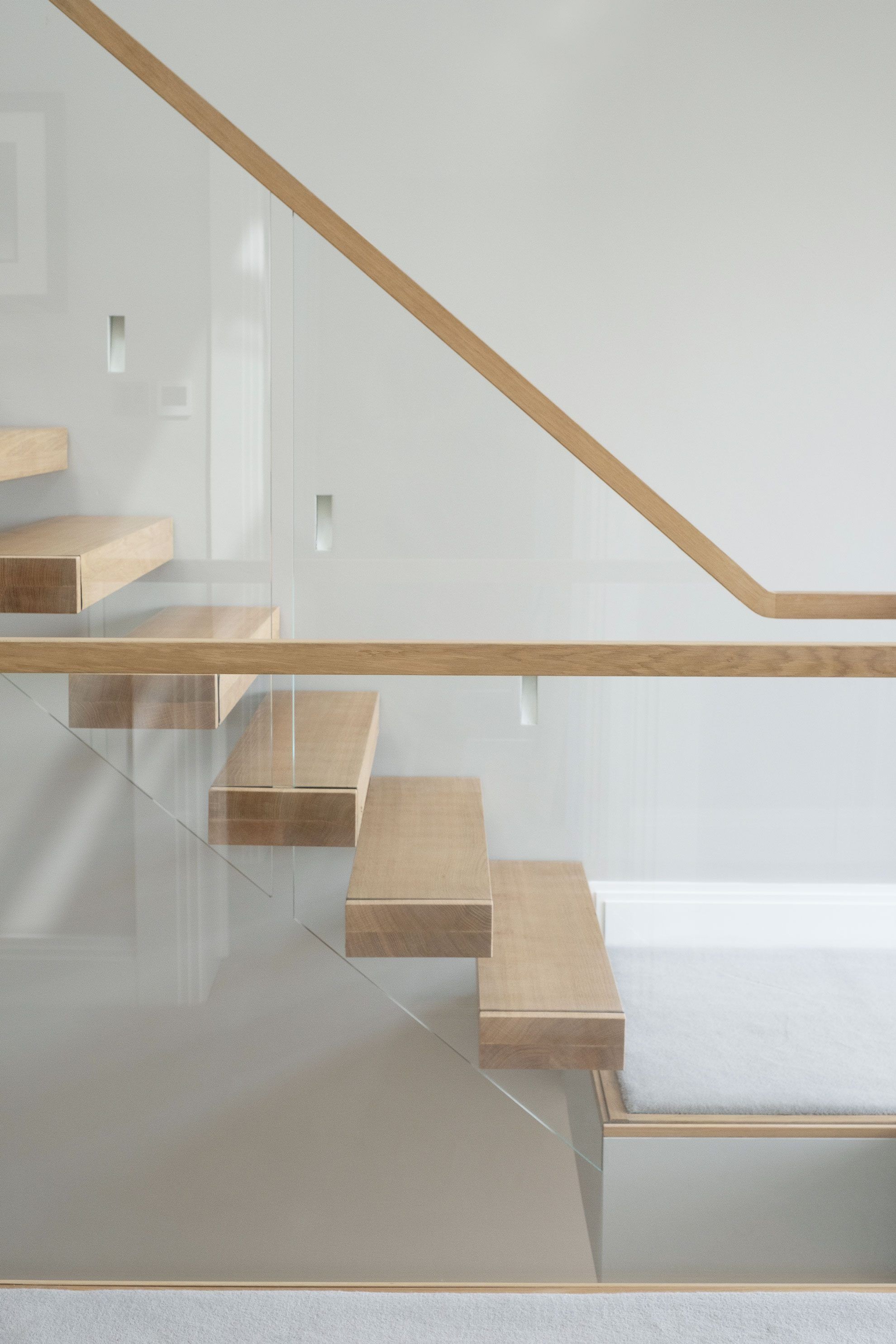 Side view of oak cantilevered floating staircase, with glass balustrade and oak handrail, with grey wall and carpet.