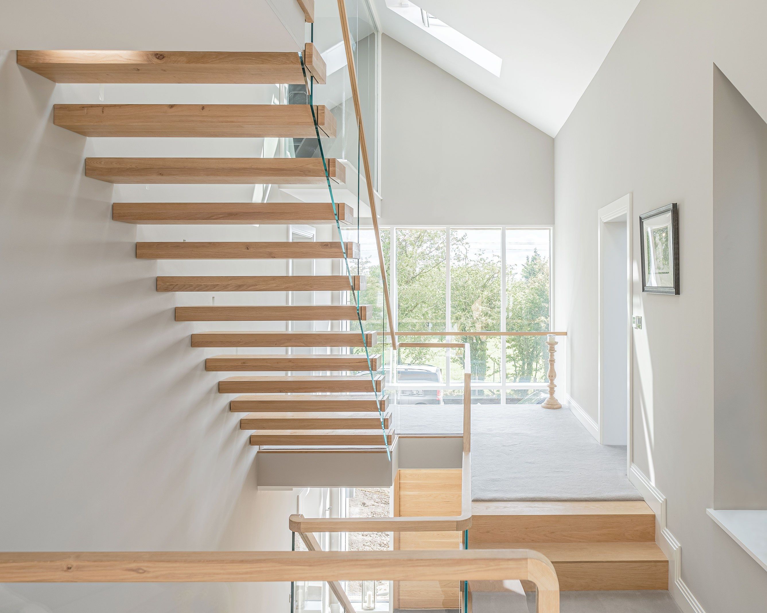 Underside view of cantilevered floating oak stair with glass balustrade from first floor. Grey walls and carpet with picture frames.