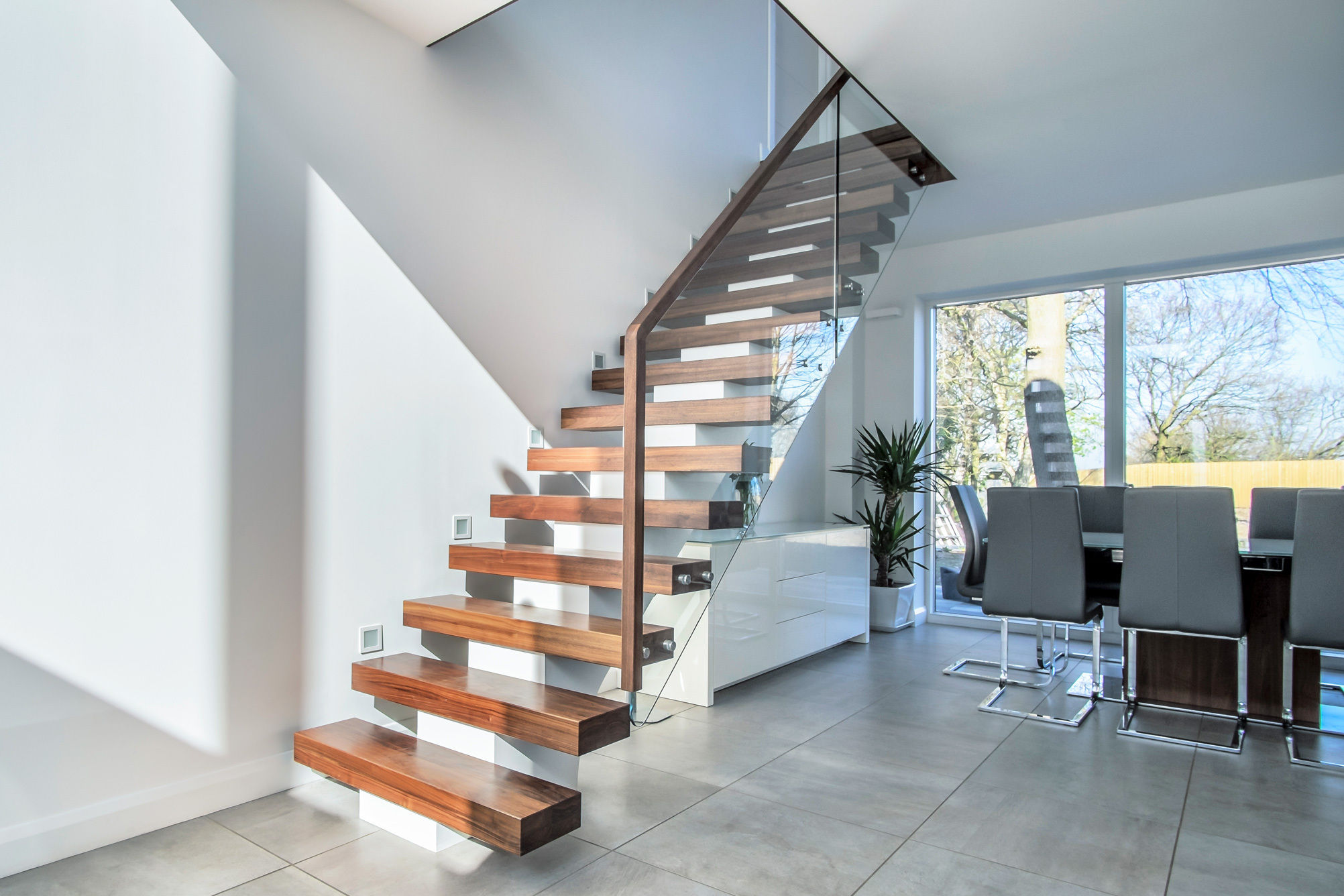 Modern domestic interior with floating central stringer feature staircase with walnut treads, handrail and glass balustrade. White walls with grey floor tiles.