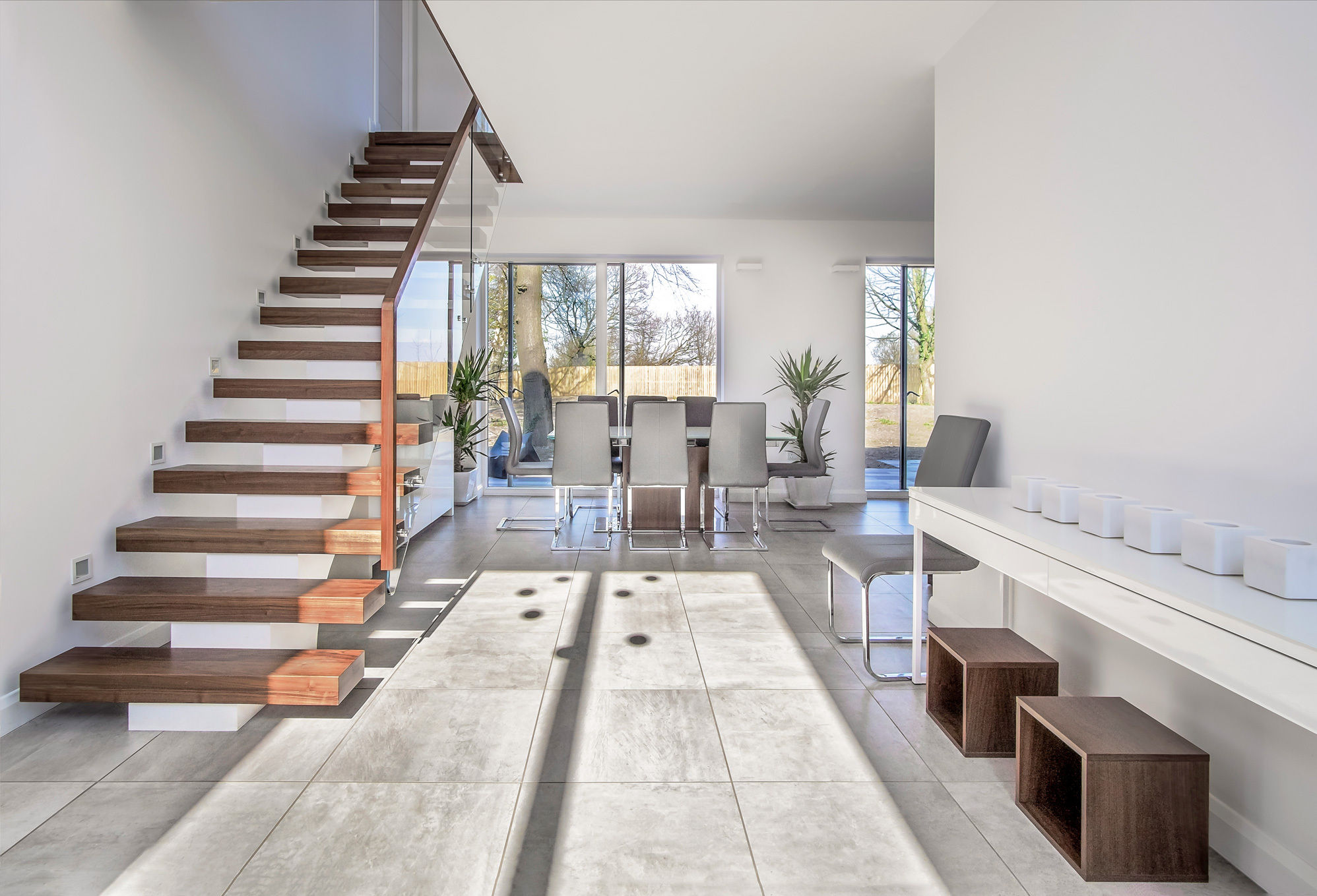 Front view of modern domestic interior with floating central stringer feature staircase. White walls with grey floor tiles and potted plants.