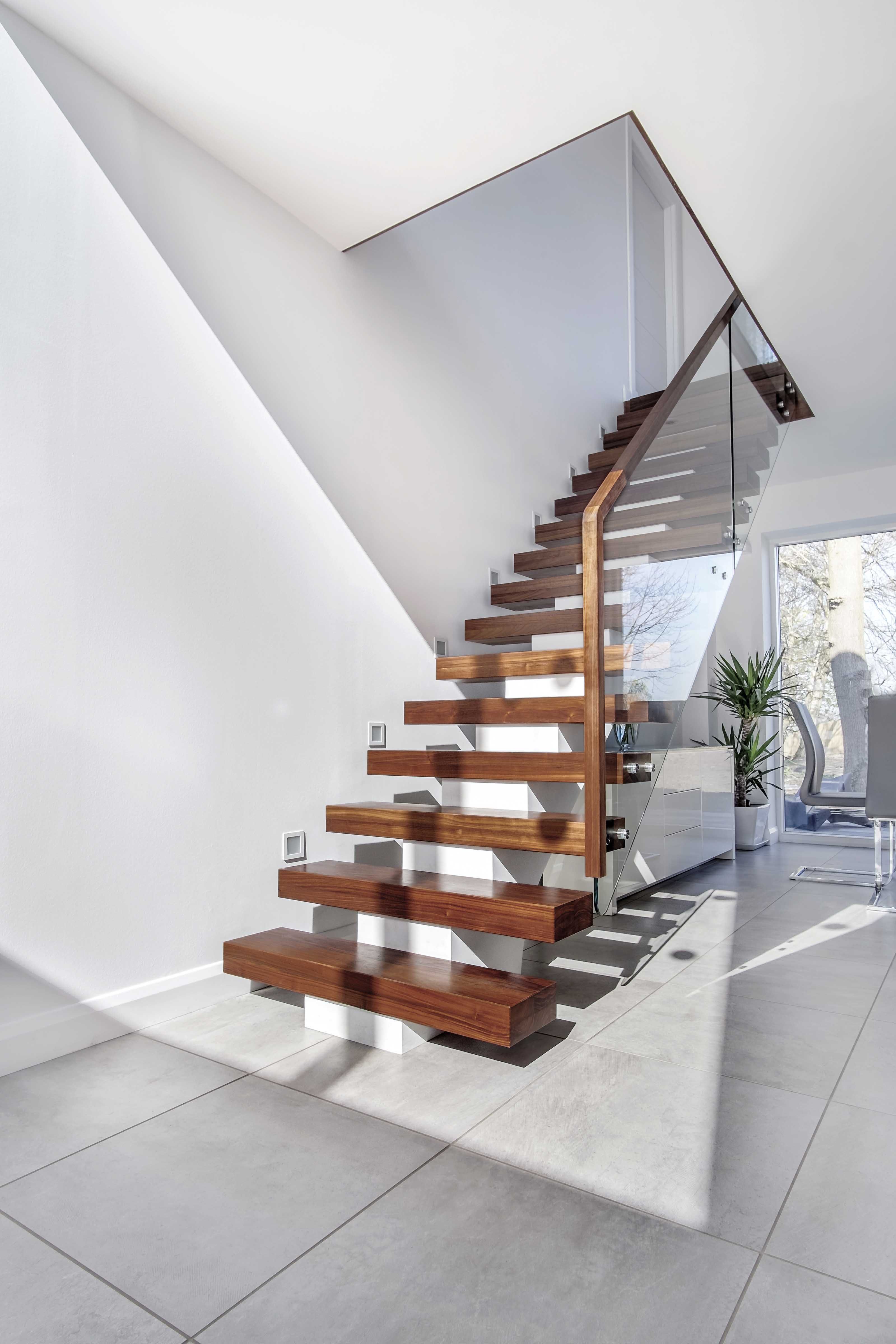 Modern domestic interior with floating central stringer feature staircase with walnut treads, handrail and glass balustrade. White walls with grey floor tiles and large potted plant.