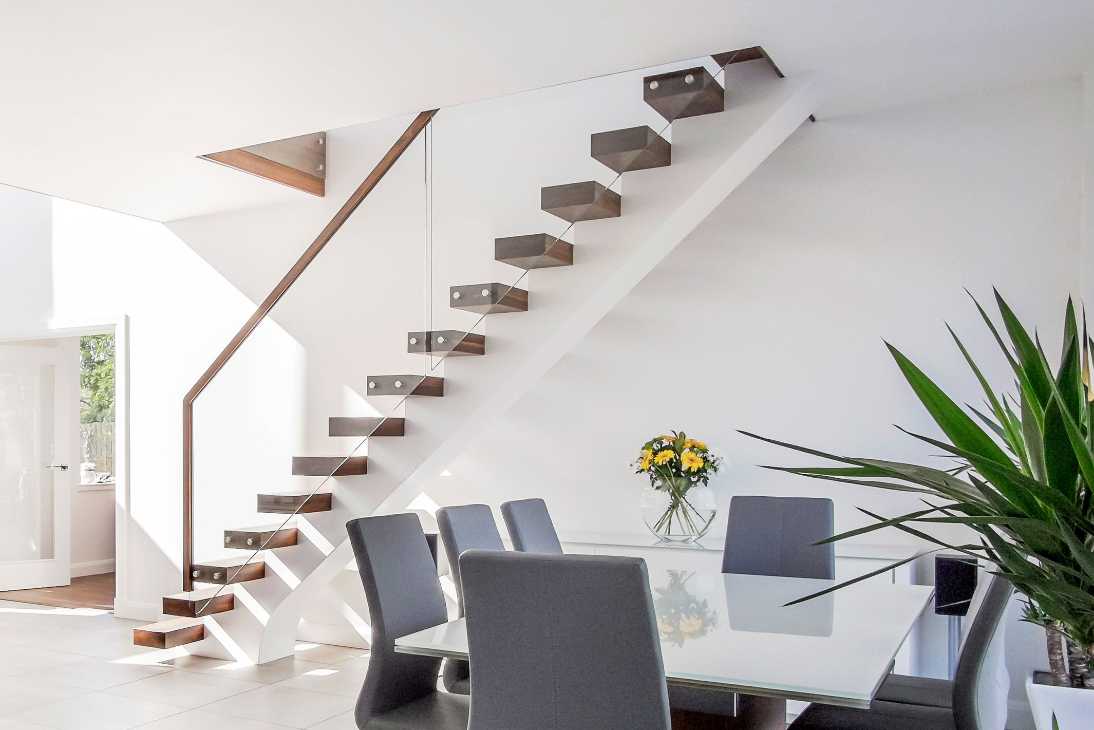 Modern domestic interior with floating central stringer feature staircase with walnut treads, handrail and glass balustrade. White walls with grey floor tiles and dining table.