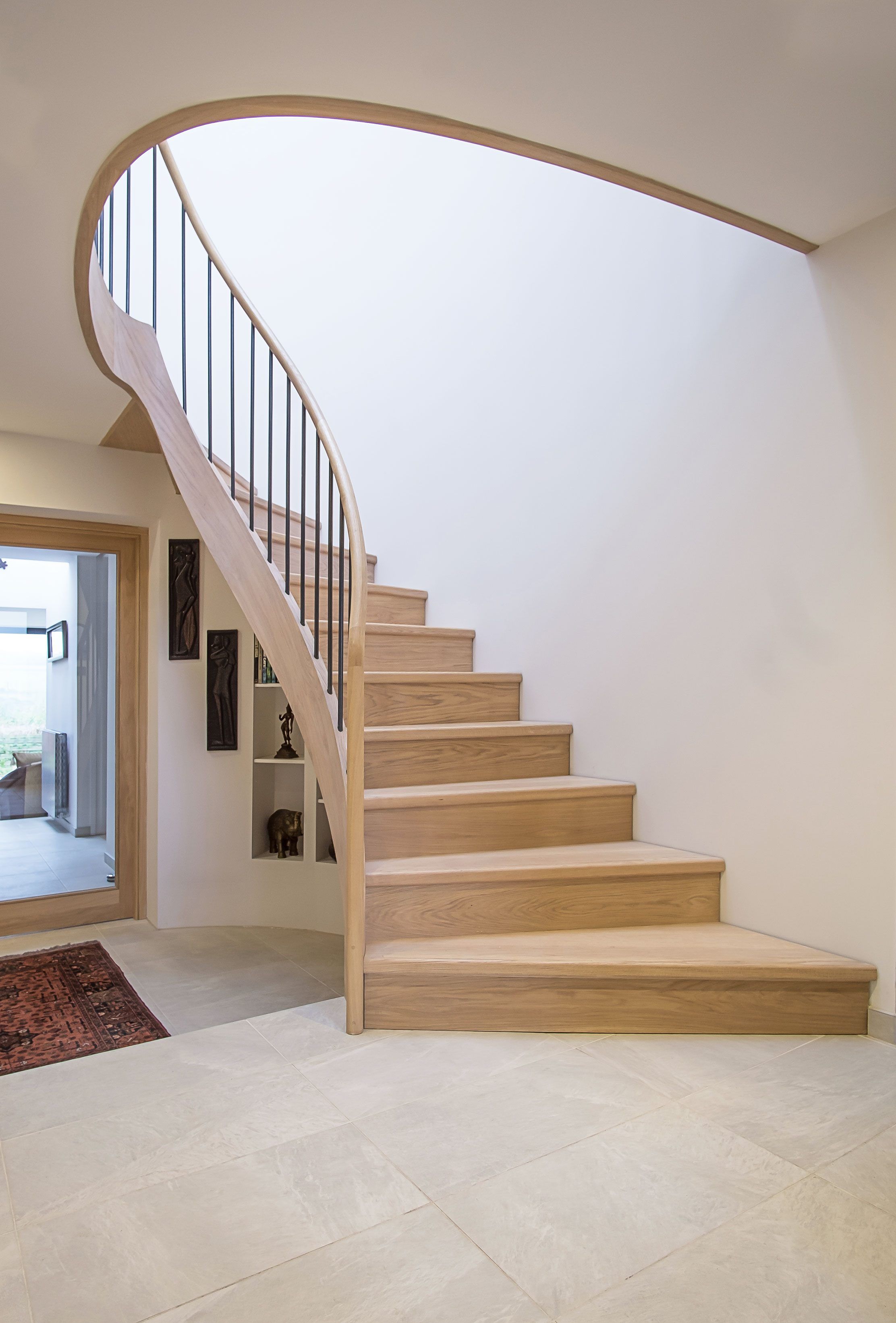 Ground floor view of feature Elliptical oak stair with iron balustrade and oak handrail. Tiled floors, rug, glazed oak doors and storage..
