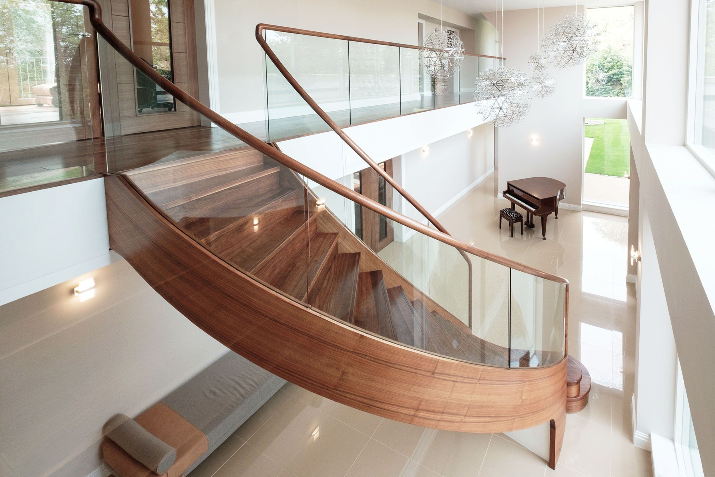 Side view of sweeping curved walnut stair with curved glass and tread lighting in large home with baby grand piano.
