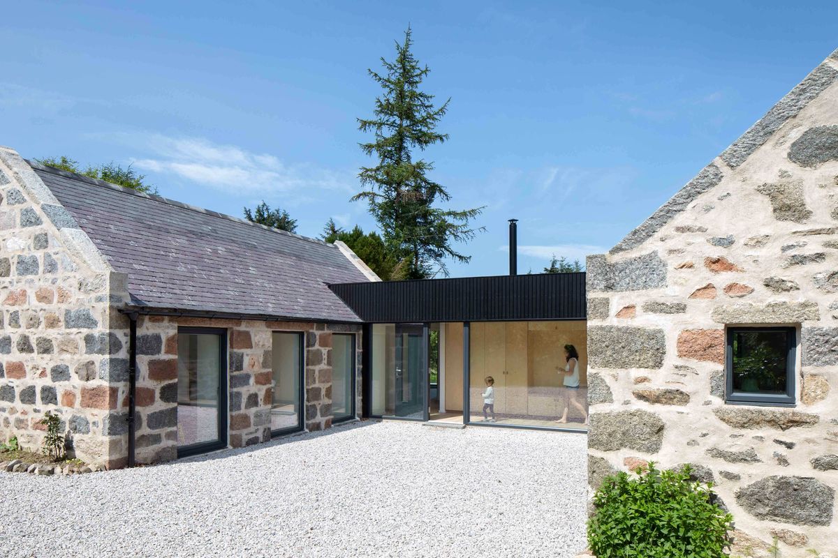 Traditional granite cottage and byre connected with modern, blackened timber and glass extension with adult and child walking.