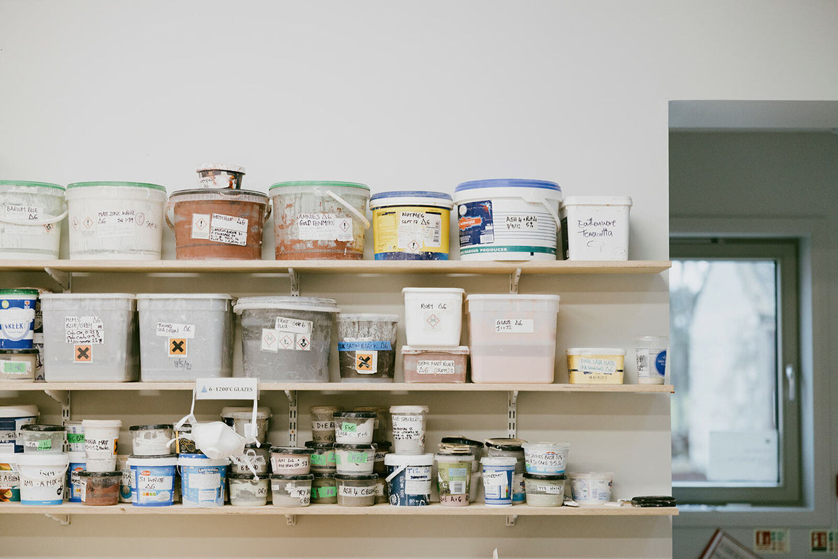 A close up photo of a 3 timber shelves. Each shelf is stacked with buckets and pots of colourful clays. The pots have handwritten labels.