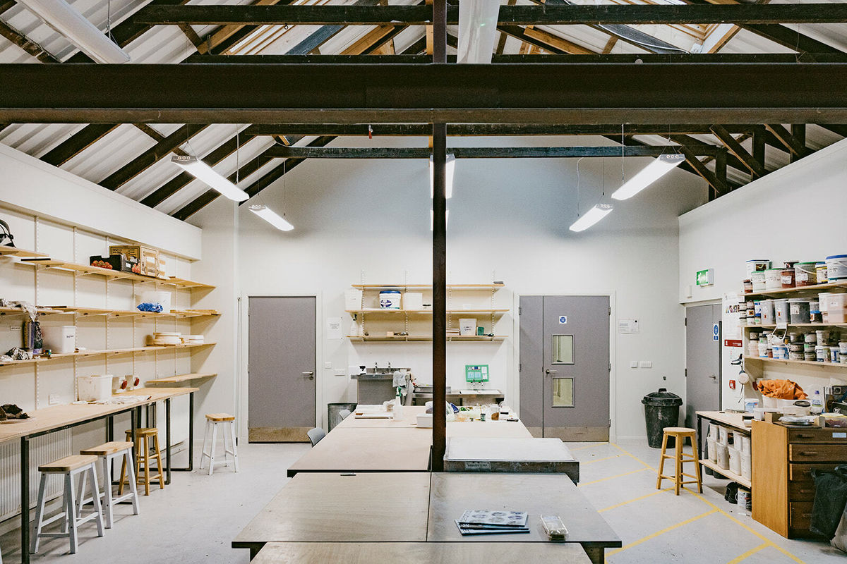 An industrial shed with exposed steel beams and white floors. The walls of the shed are lined with shelves. On the shelves sit tins of paint and buckets of plaster. Stools are dotted around the shed.