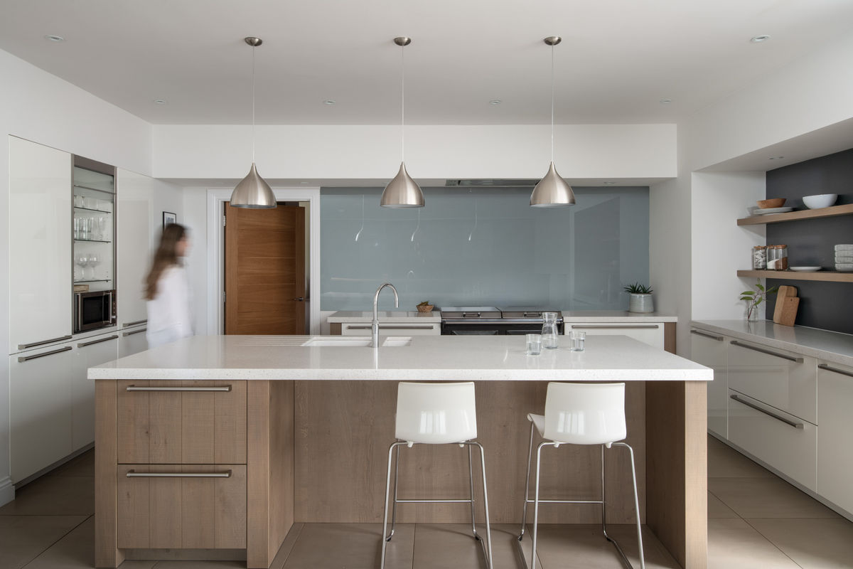 A woman walks by a kitchen island with white marble countertops and timber cabinetry. Three grey downlights hang over the kitchen island. A grey glass splashback is on the back wall, sitting above a range cooker.