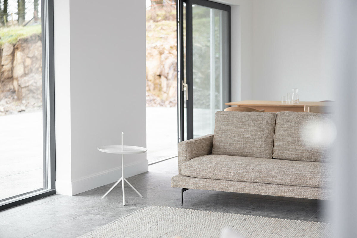 A living room setting. With a couch and small white side table in the foreground and a wooden dining table in the background. The walls are painted white.