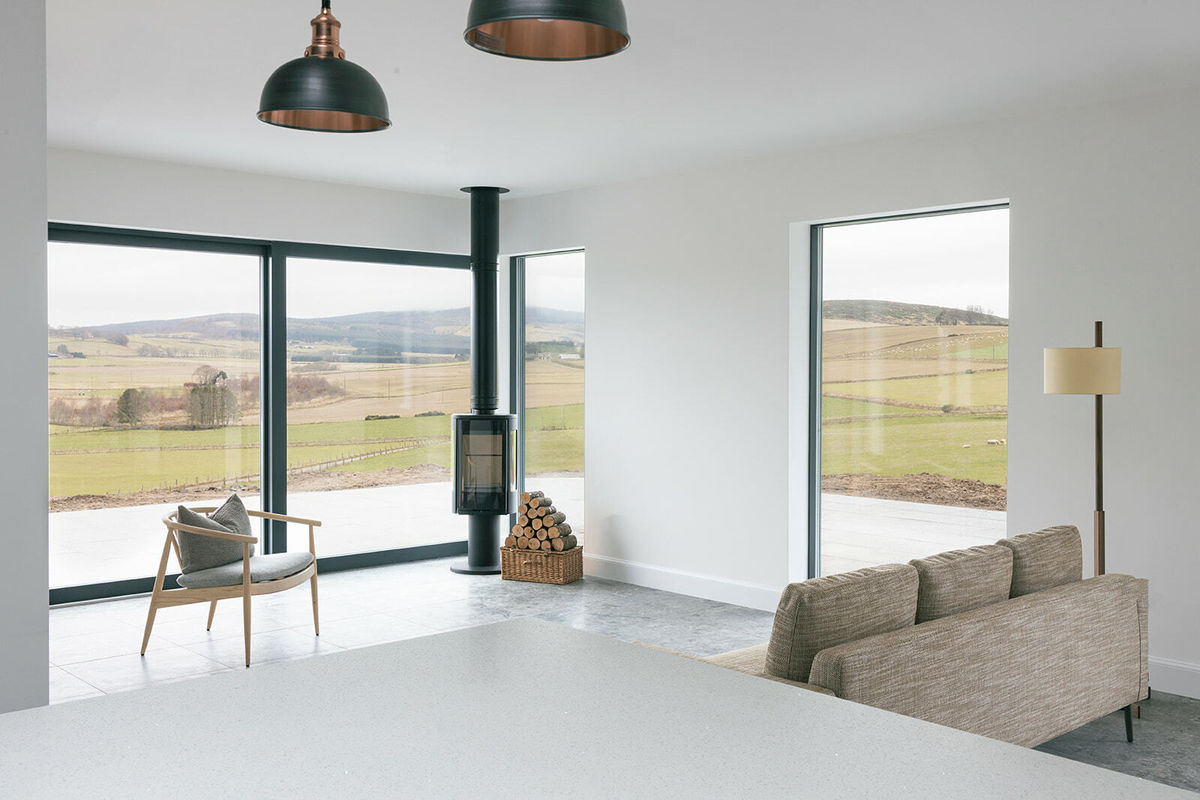 A living room with a panoramic rural view, a couch and a chair. A black wood burning stove in the corner of the room with logs next to it.