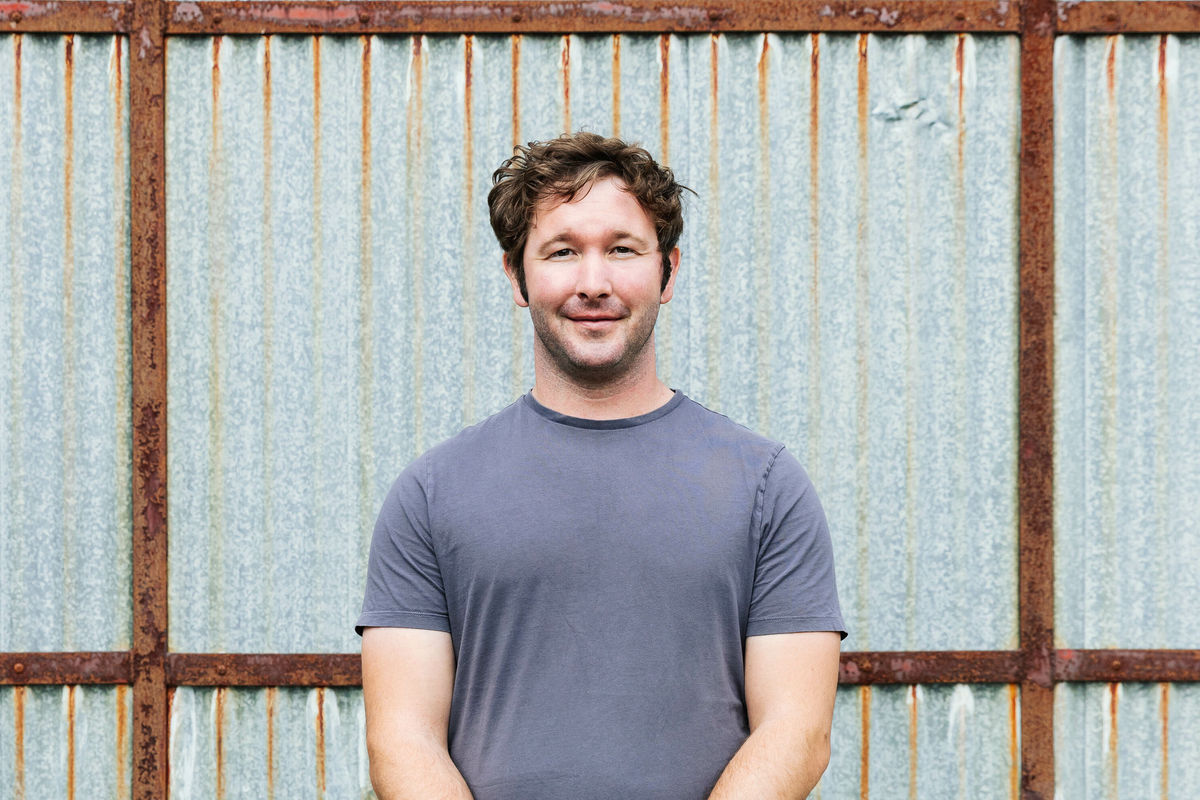 A man in a blue shirt stands in front of a large, rusty, corrugated iron door. He smiles.