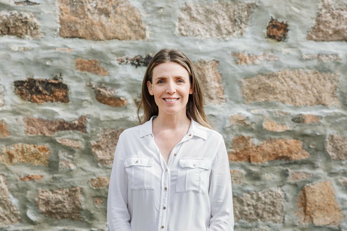 A woman in a white shirt stands in front of a granite stone wall. She smiles.