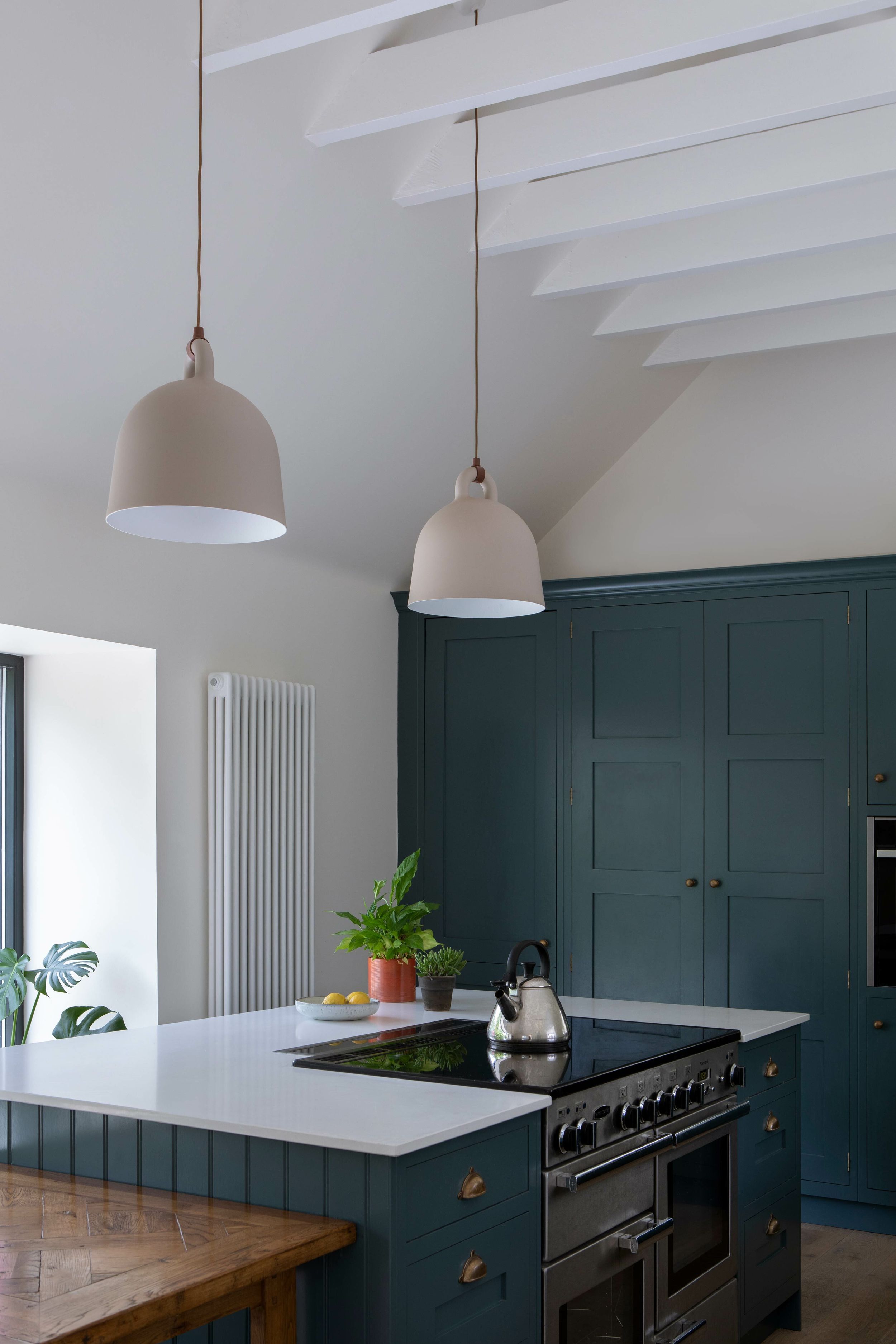 A kitchen with exposed ceiling ties, dark blue cabinetry and a timber table. A kettle sits on top of a black hob.