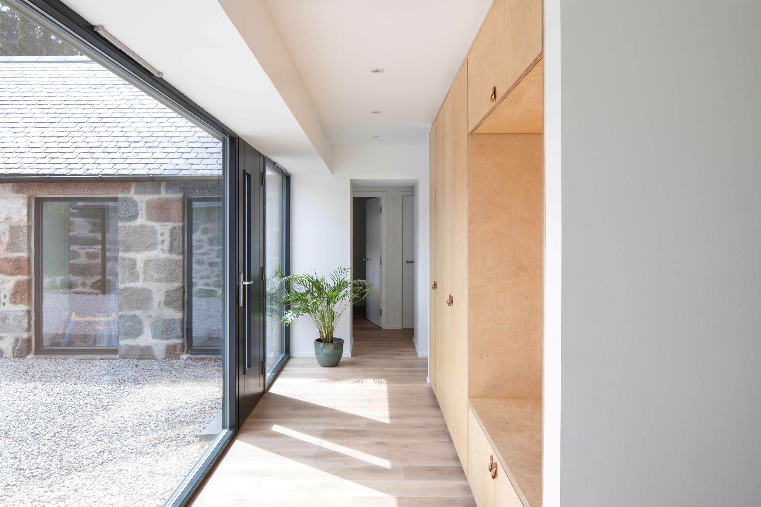 Birch cabinetry runs along the right hand side of a hallway. The cabinetry has a built in bench to sit. Floor to ceiling glazing is on the left hand side of the hallway, with a door in the centre.