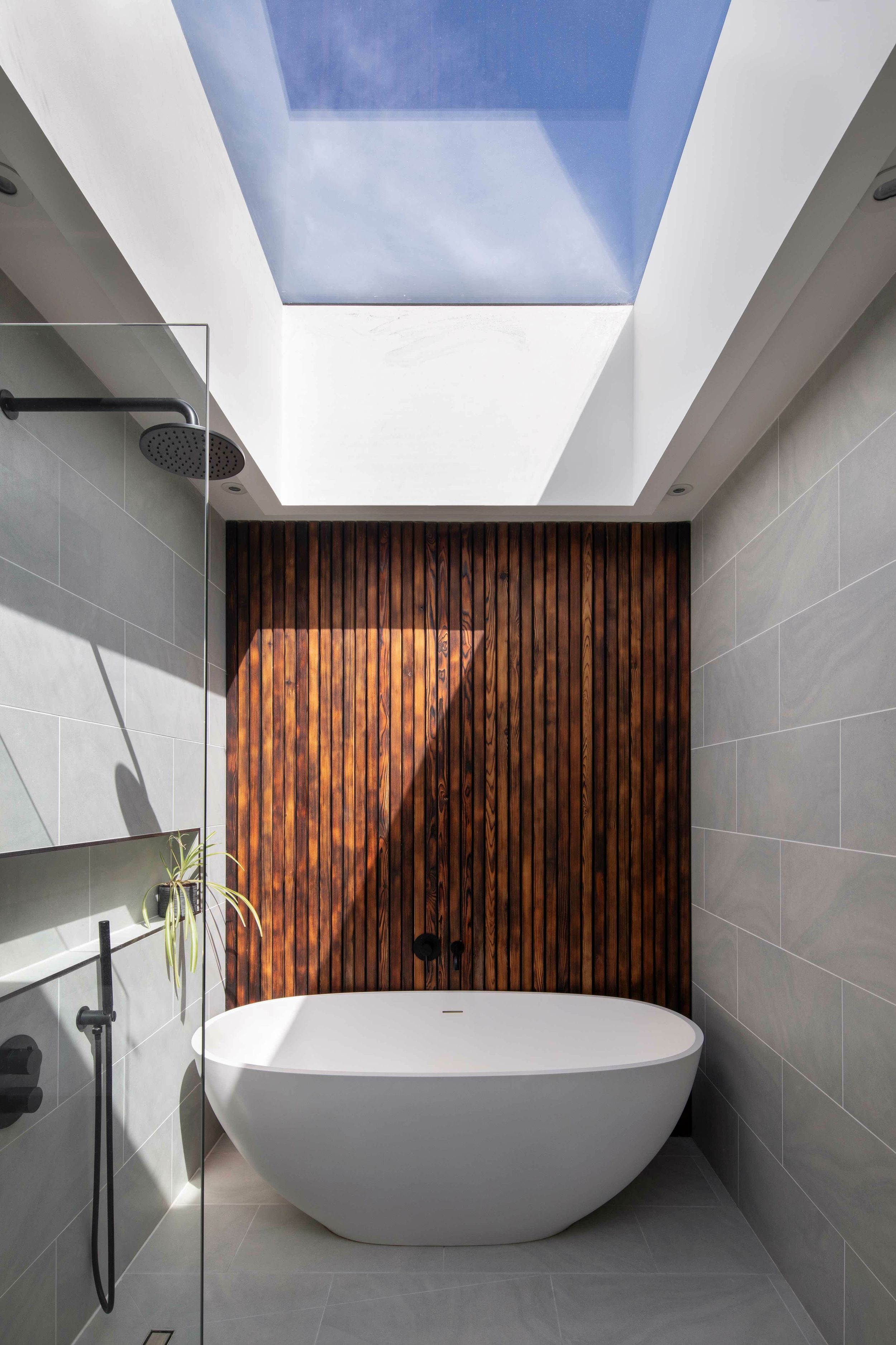 A skylight lets sunlight into a grey-tiled bathroom, illuminating a walk-in shower and a white bath. Behind the bath is a feature wall made from charred larch timber.