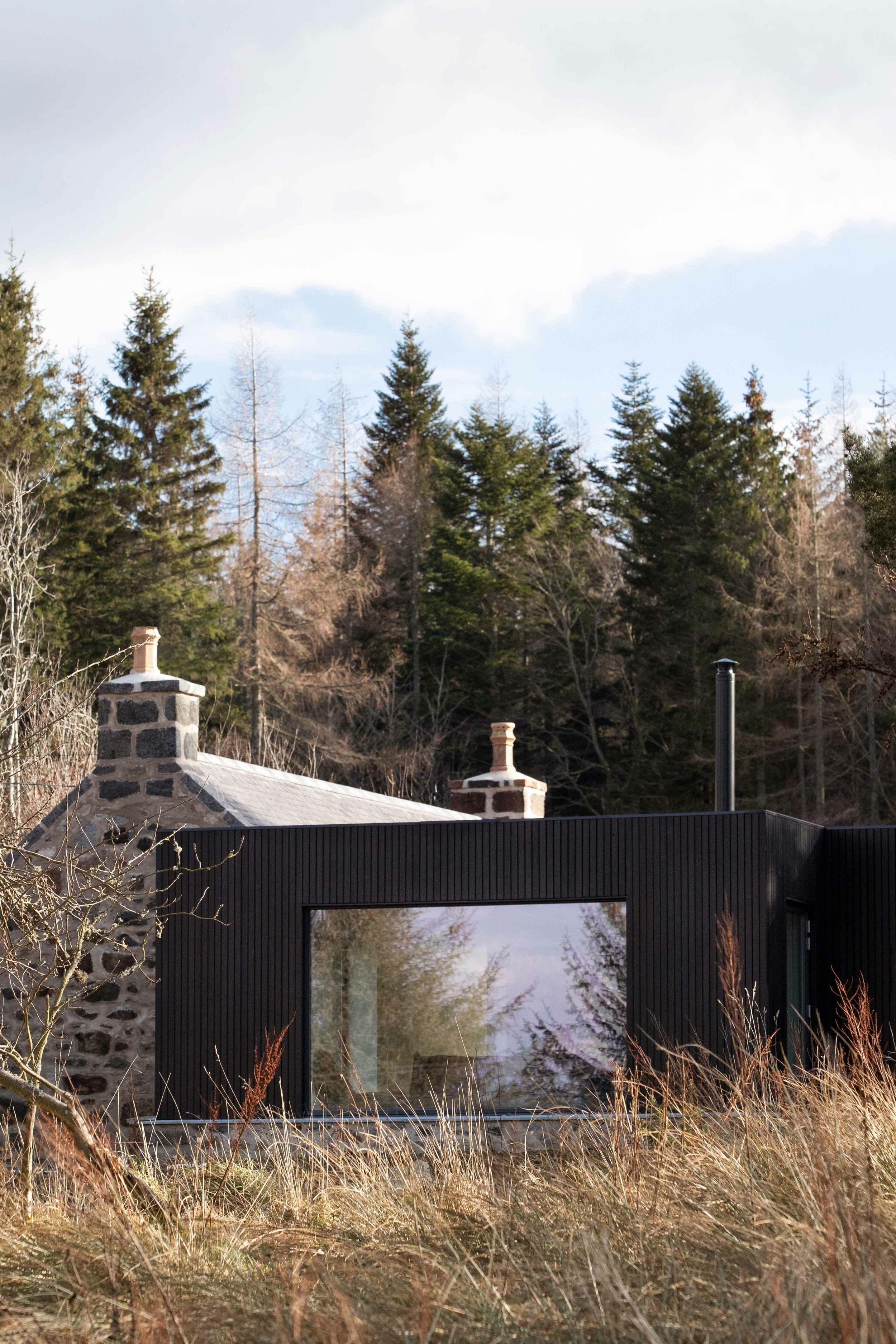 Modern, blackened timber and glass box shaped extension and granite cottage, surrounded by woodland.