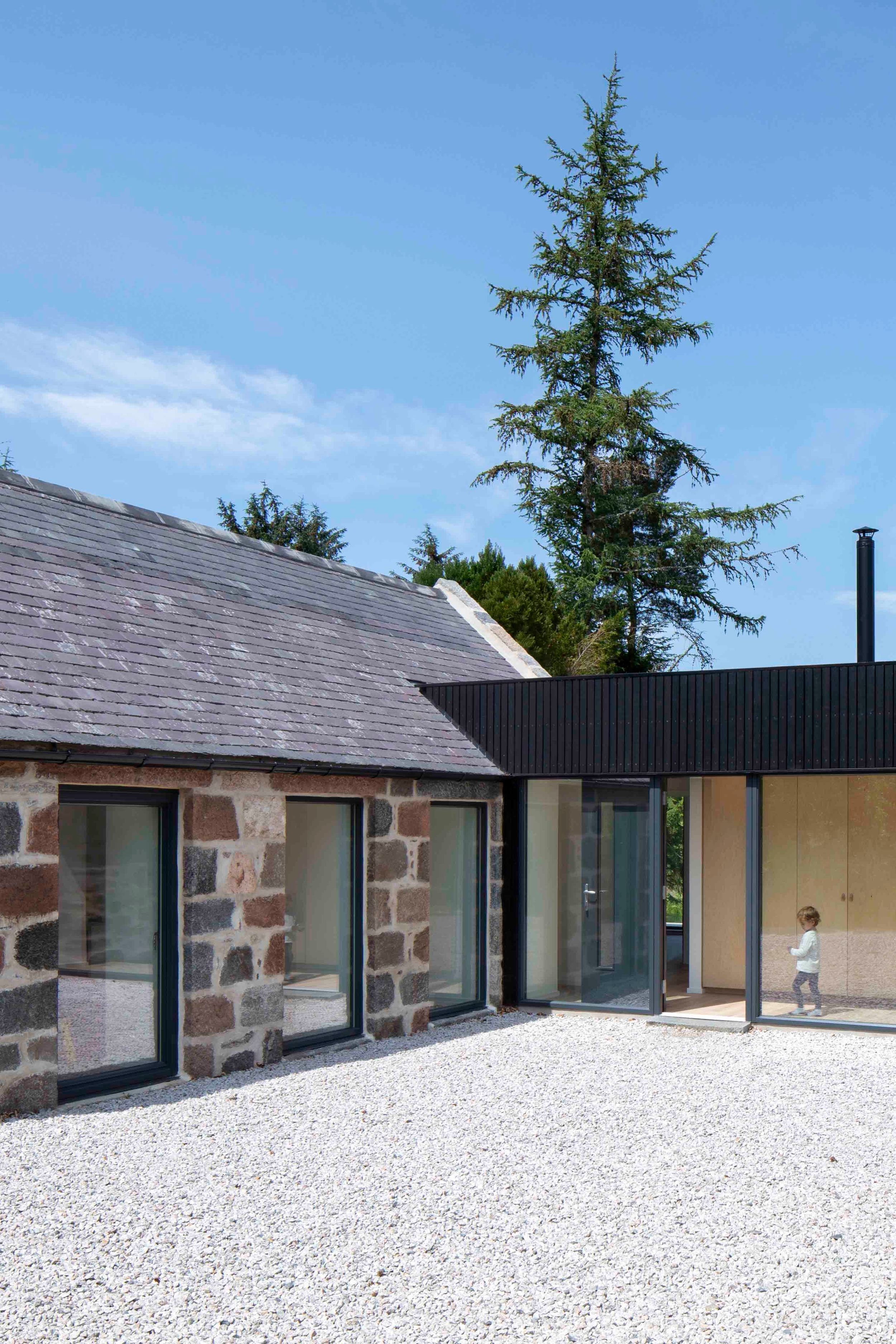 Traditional granite cottage and byre connected with modern, blackened timber and glass extension with a child walking.