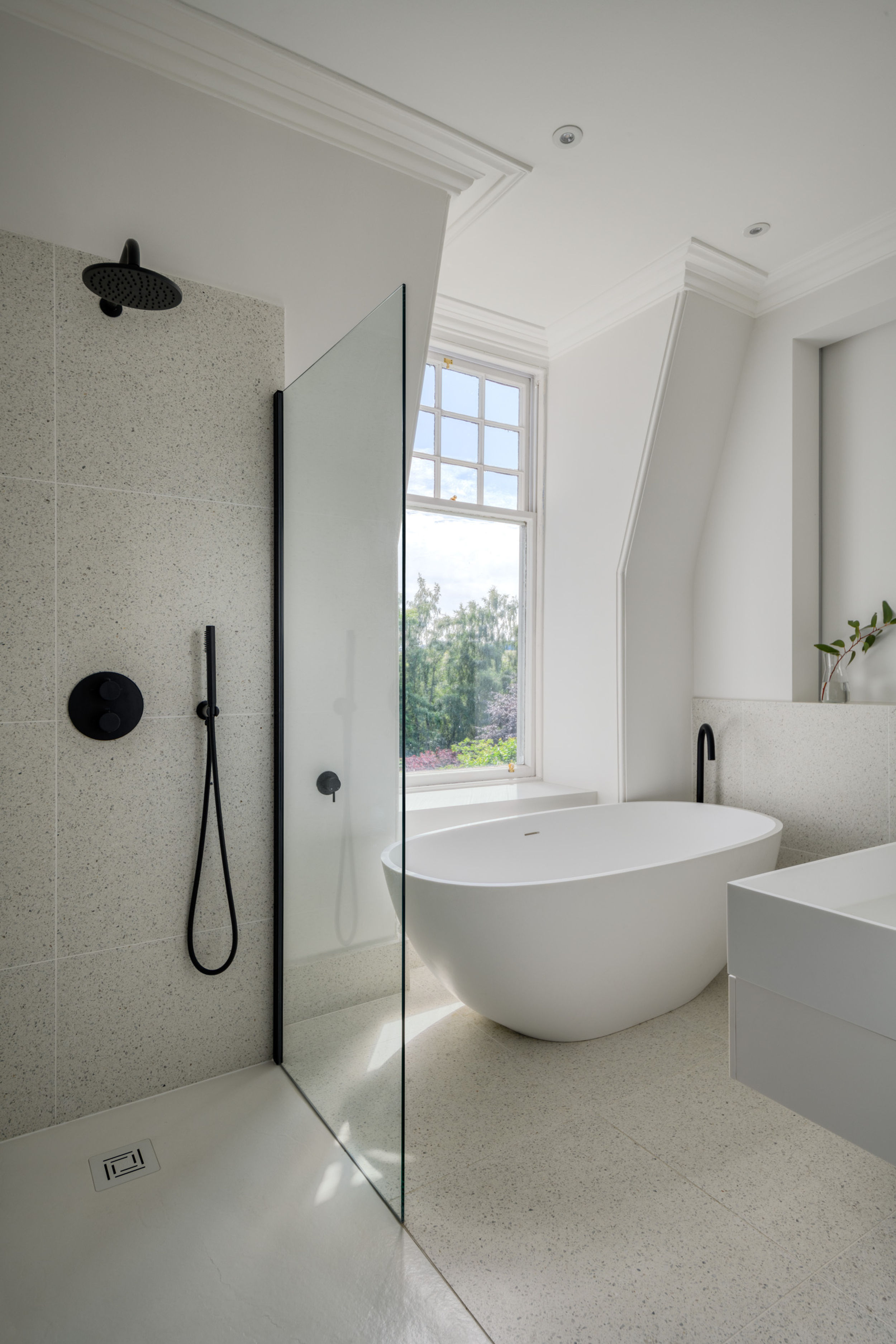 Tiled bathroom with stand-up shower and black fixtures, a white freestanding bath and window view of trees