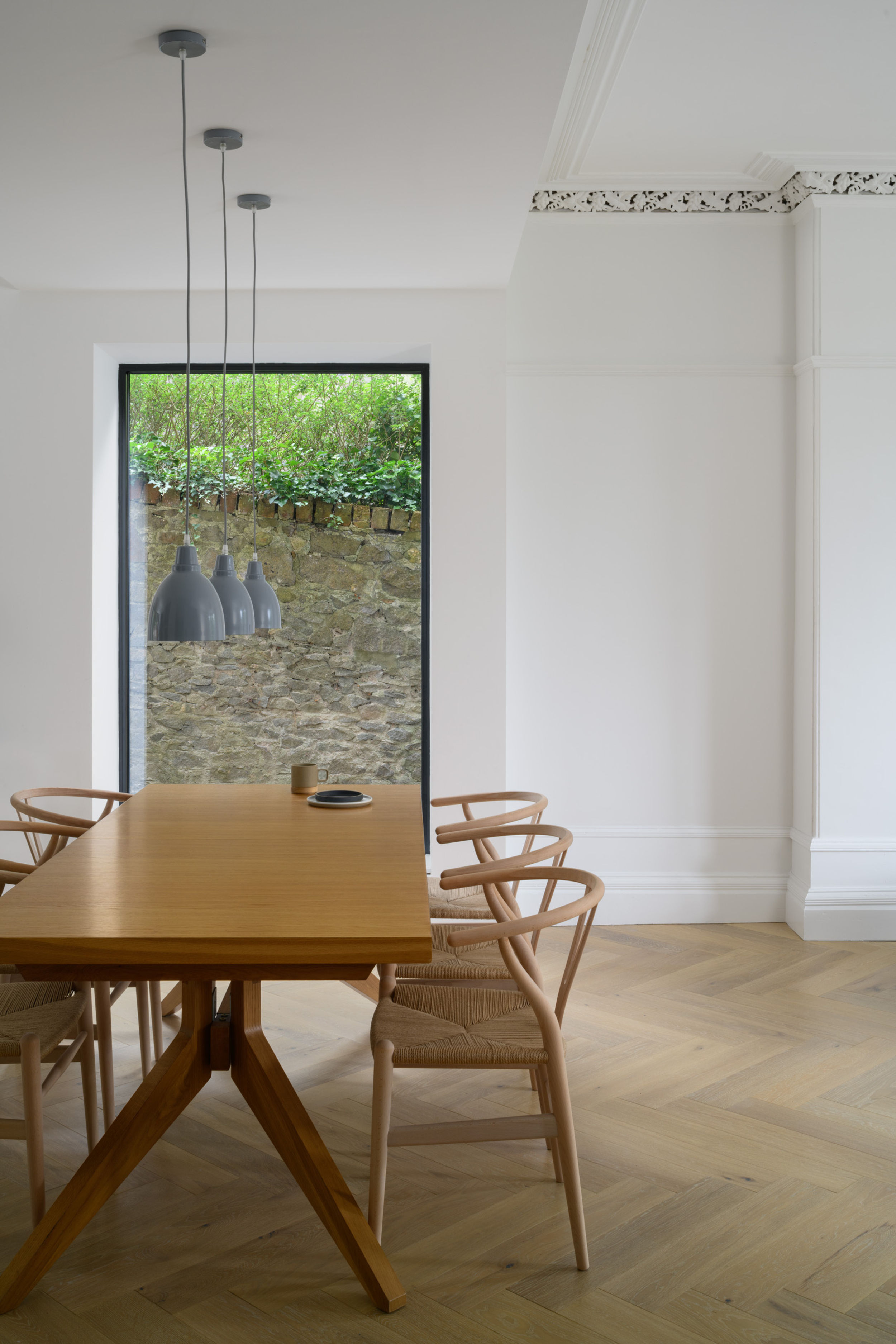 Large wooden dining table with five wishbone chairs, grey hanging lamps and tall vertical window