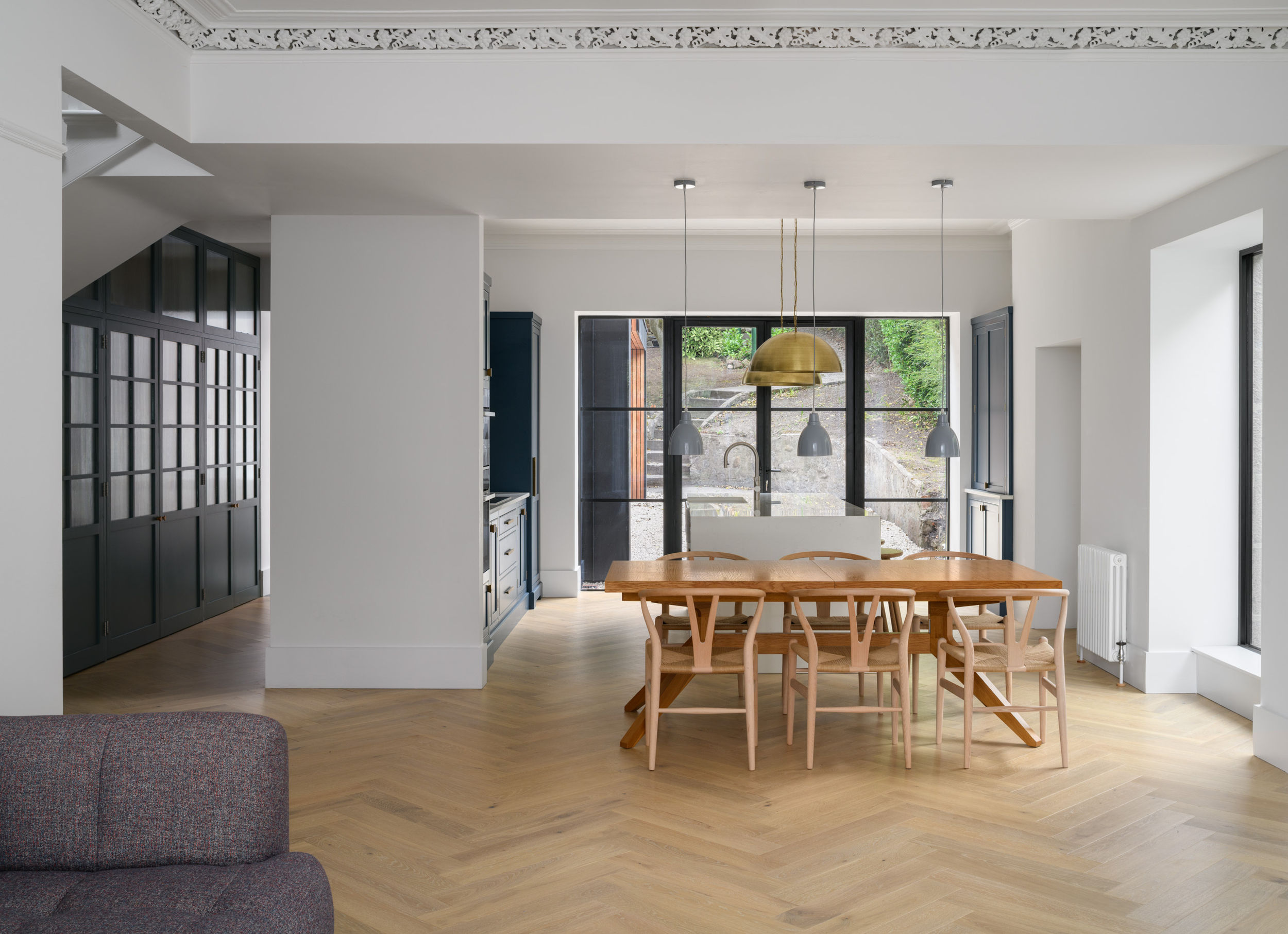 Large family-style kitchen, hallway and dining area with wooden table and six wishbone chairs. White walls, herringbone flooring and hanging lights. View to garden through Crittal style doors.