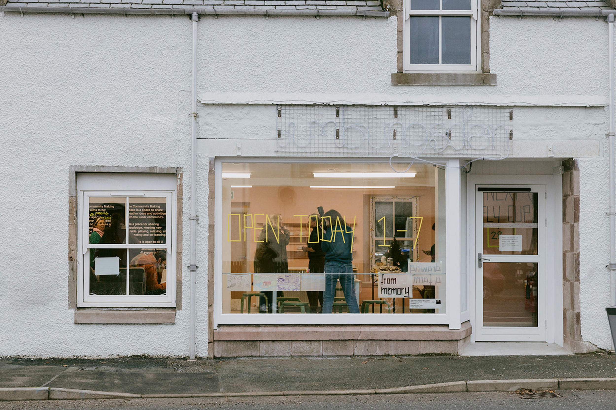 An exterior of a shopfront with two windows, a large and small. There is a door on the left hand side. Through the window you can see people standing inside the shop.