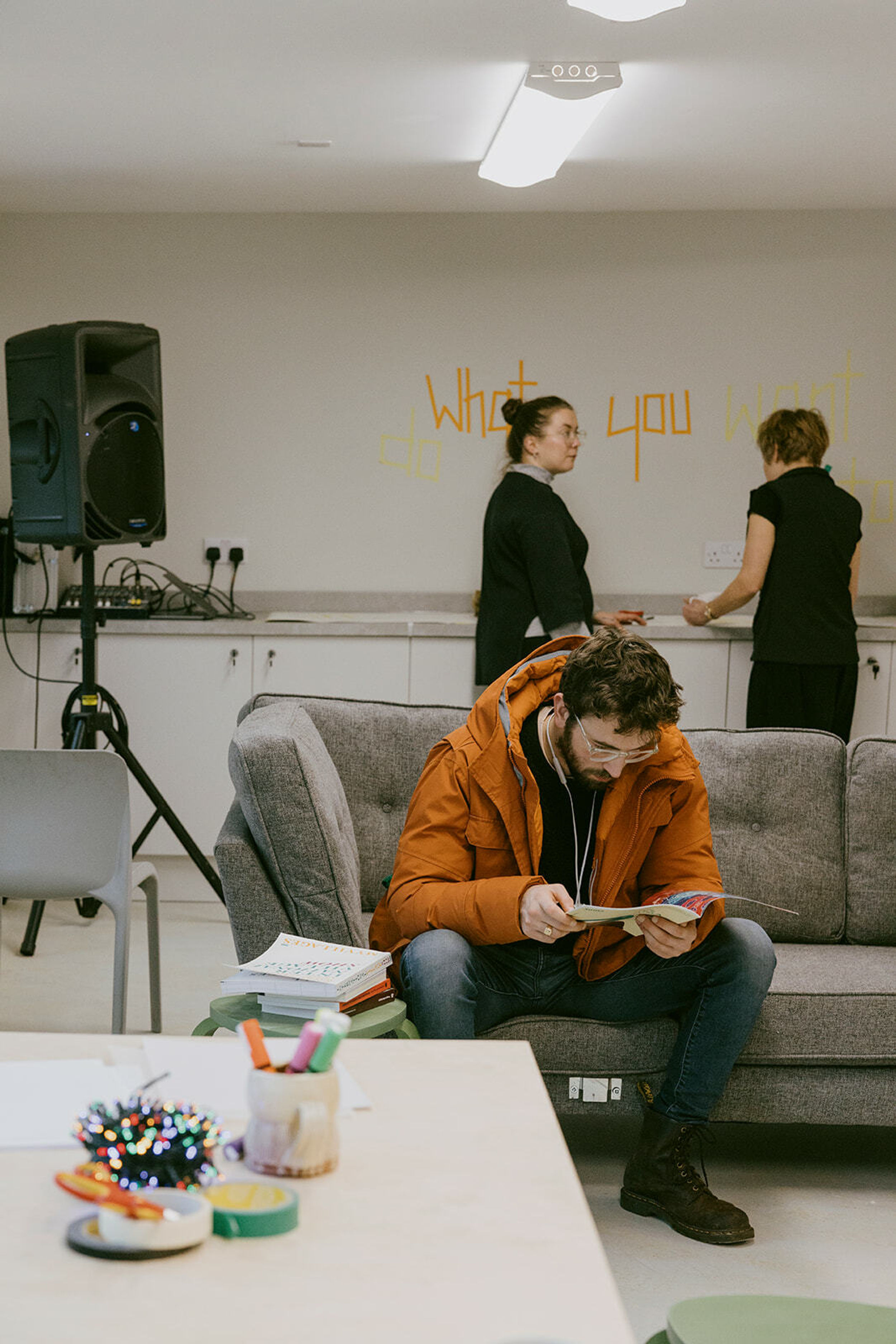 A person wearing an orange jacket sits on a couch reading a magazine. Two people in the background are having a conversation and leaning against a worktop.