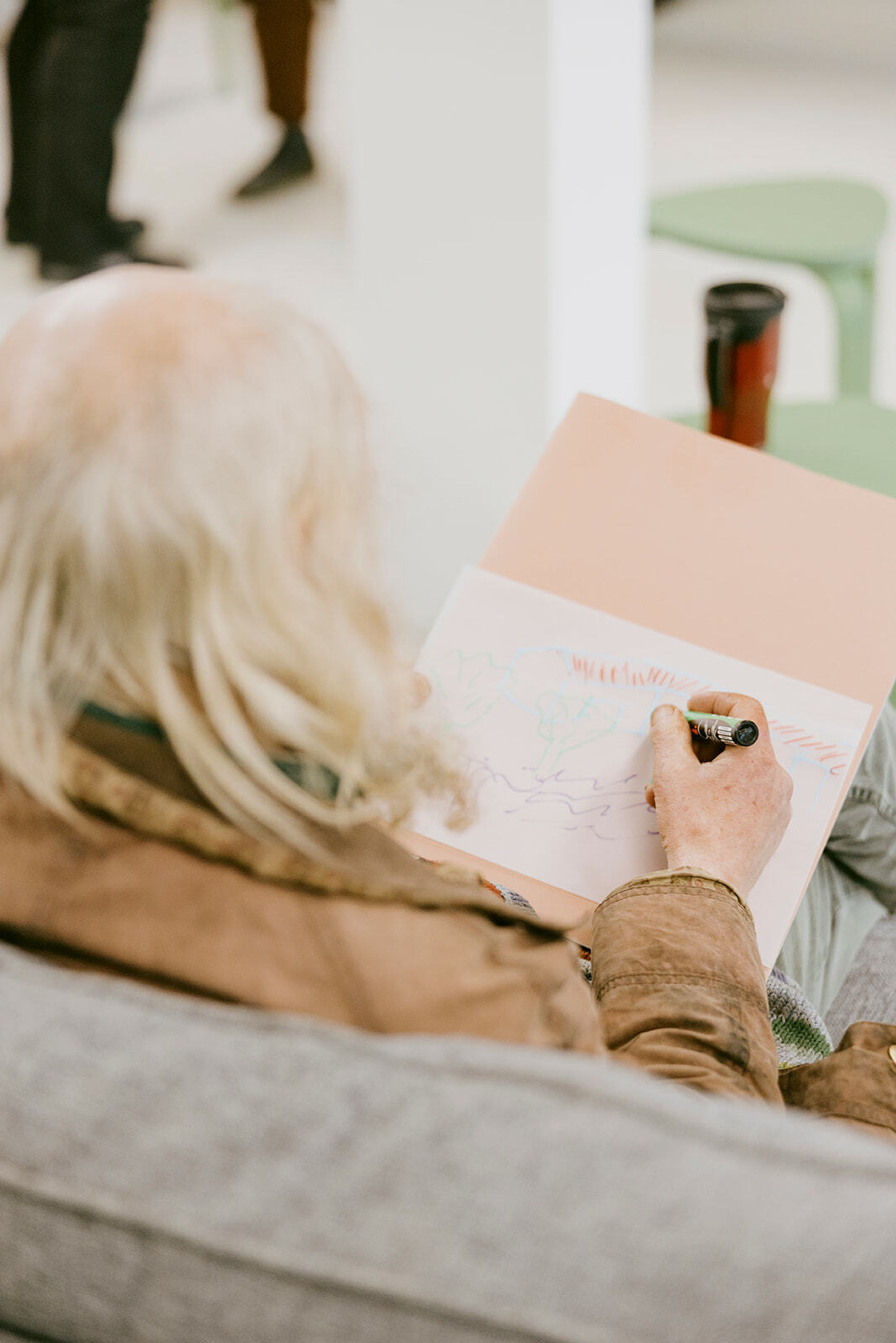 Looking over the shoulder of a person with white, whispy hair. They hold a pen in their right hand and are drawing on paper.