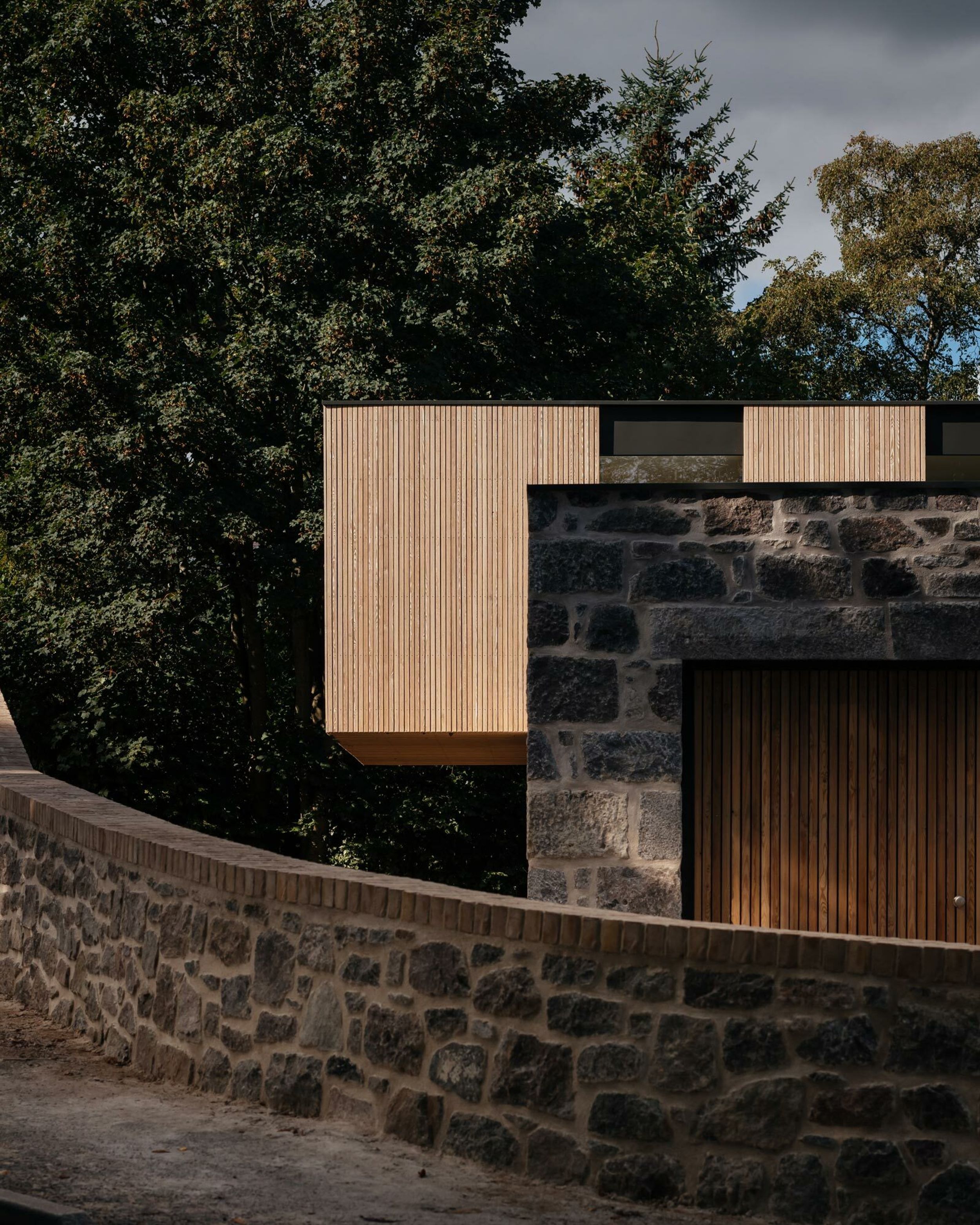 A cantilevered, timber clad house. A granite stone boundary wall surrounds the property.