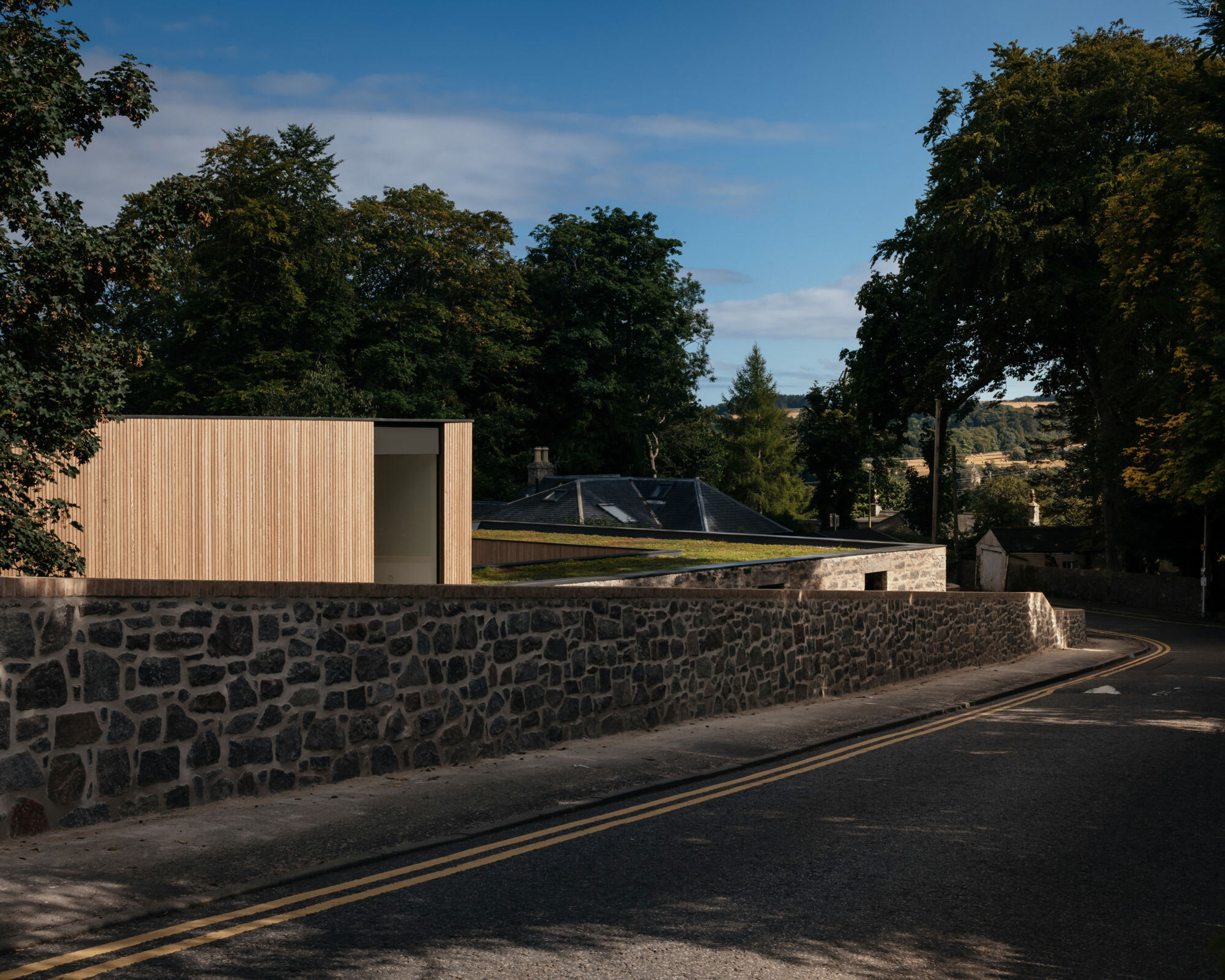 An external view looking at a contemporary, natural timber clad home from across a road. A stone wall encloses the home, which has a flat green sedum roof.