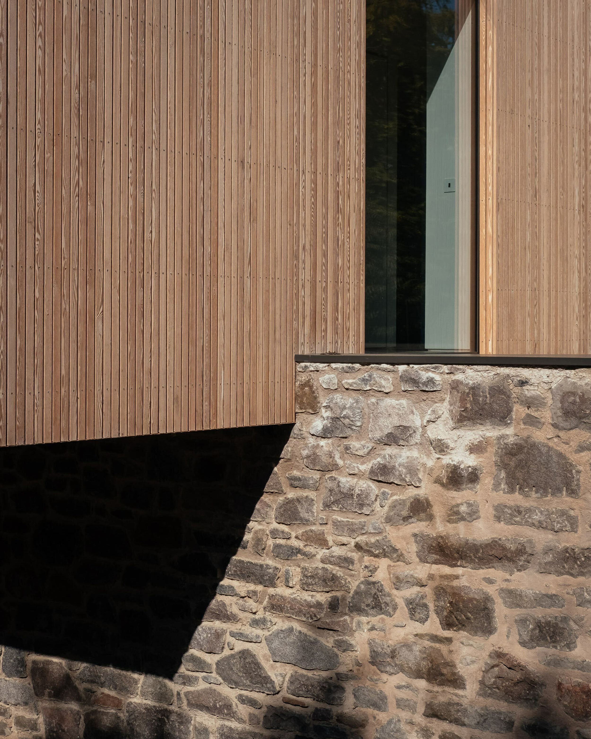 A close up of a granite stone wall. A modern house, clad in timber cuts into the wall and casts a shadow on the stone.