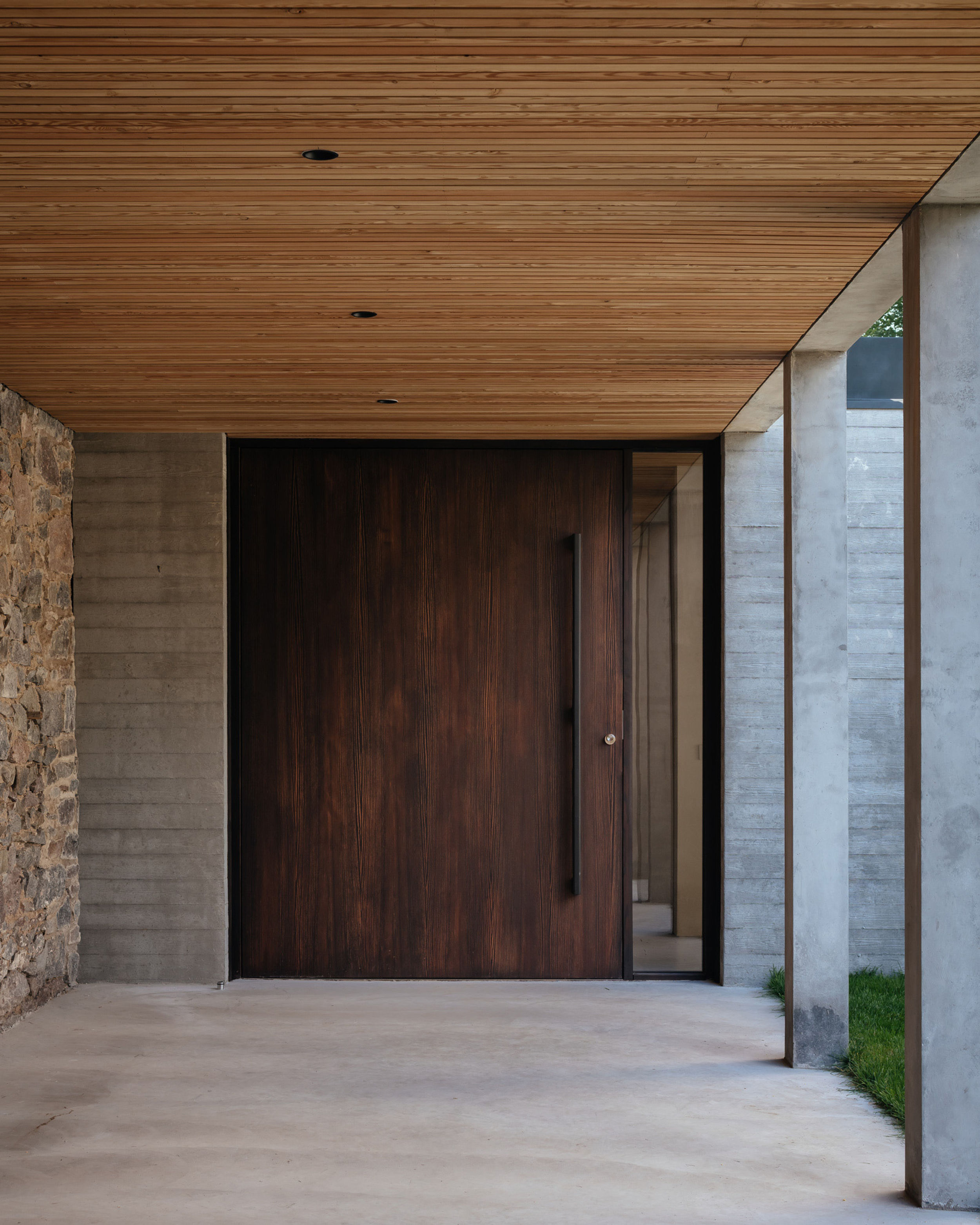 Looking down a concrete colonnade with a timber lined ceiling. One wall of the colonnade has been made with an old stone wall. A smoked larch pivot front door is at the end of the colonnade.