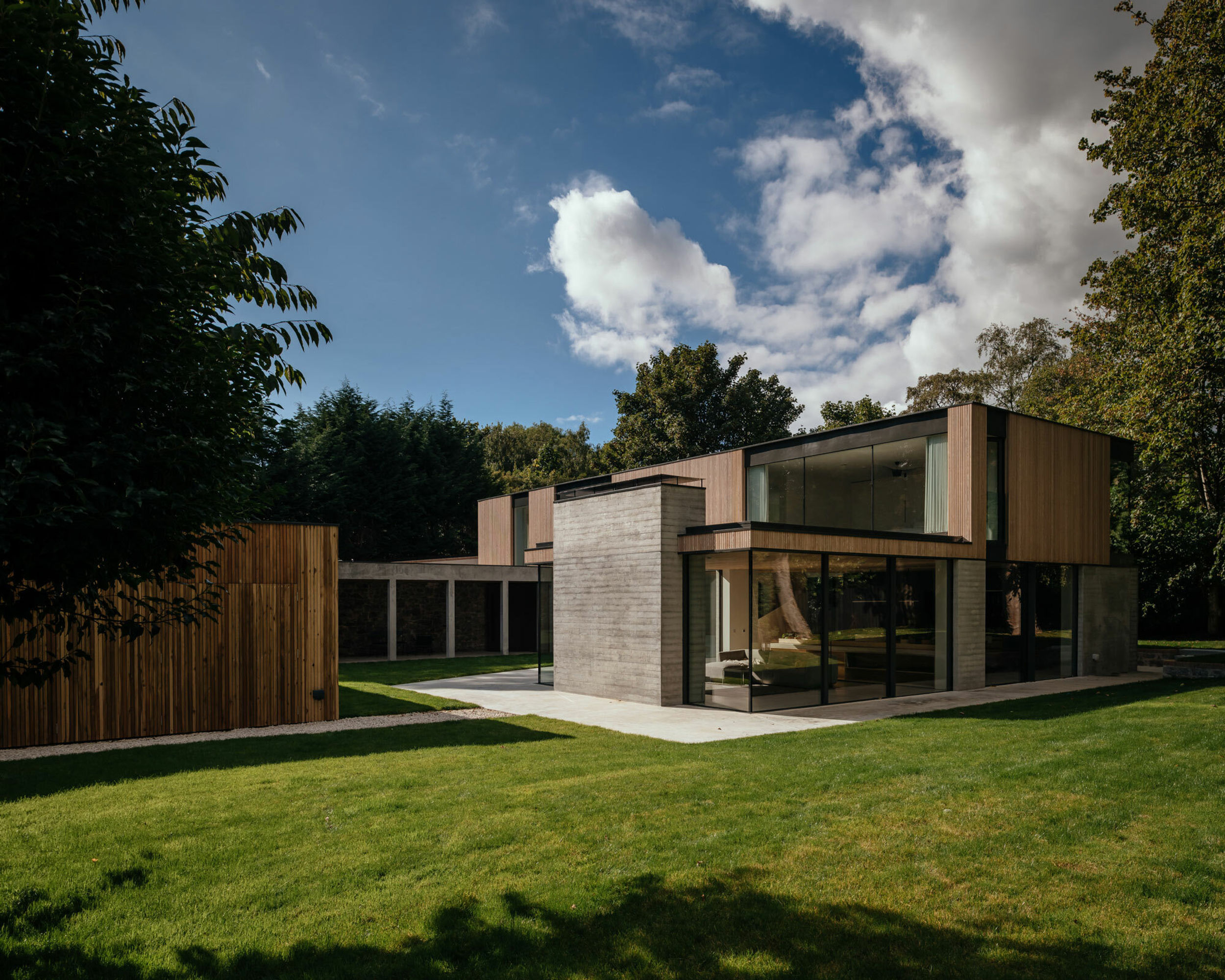 A wide shot of a modern home. The first floor- a long, elevated, timber and glass building- cantilevers above a concrete colonnade. There is lots of floor to ceiling glass in the property and you can see right through to the other side. The house is made from timber cladding and board marked concrete. Trees and garden surround the property.