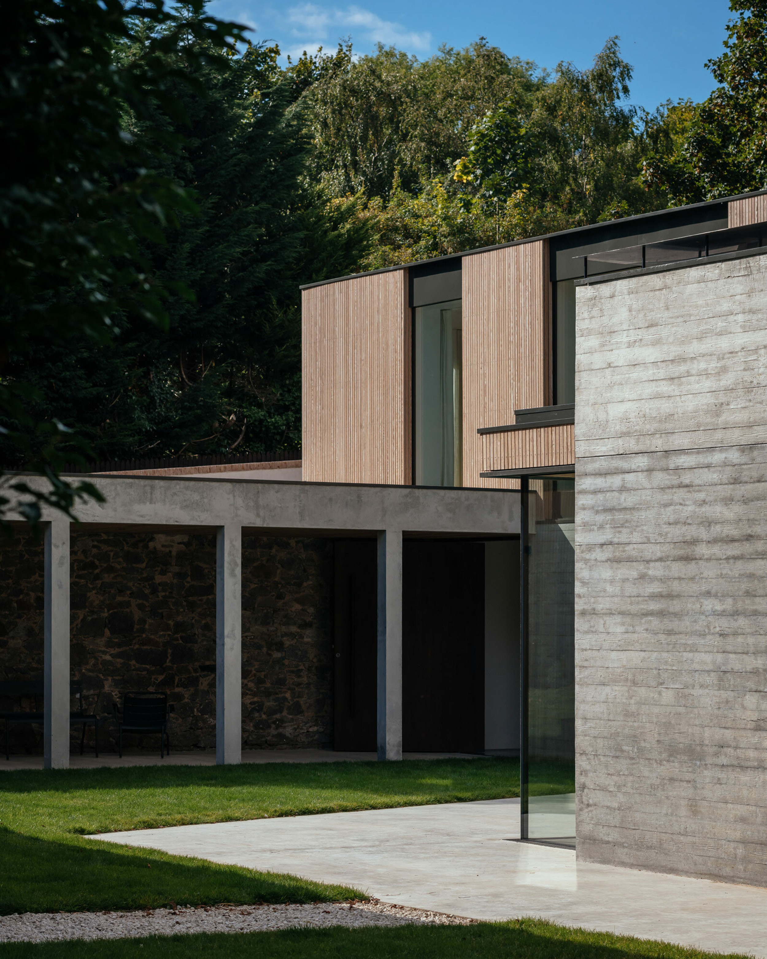 A wide shot of a modern home. The first floor- a long, elevated, timber and glass building- cantilevers above a concrete colonnade. There is lots of floor to ceiling glass in the property and you can see right through to the other side. The house is made from timber cladding and board marked concrete. Trees and garden surround the property.