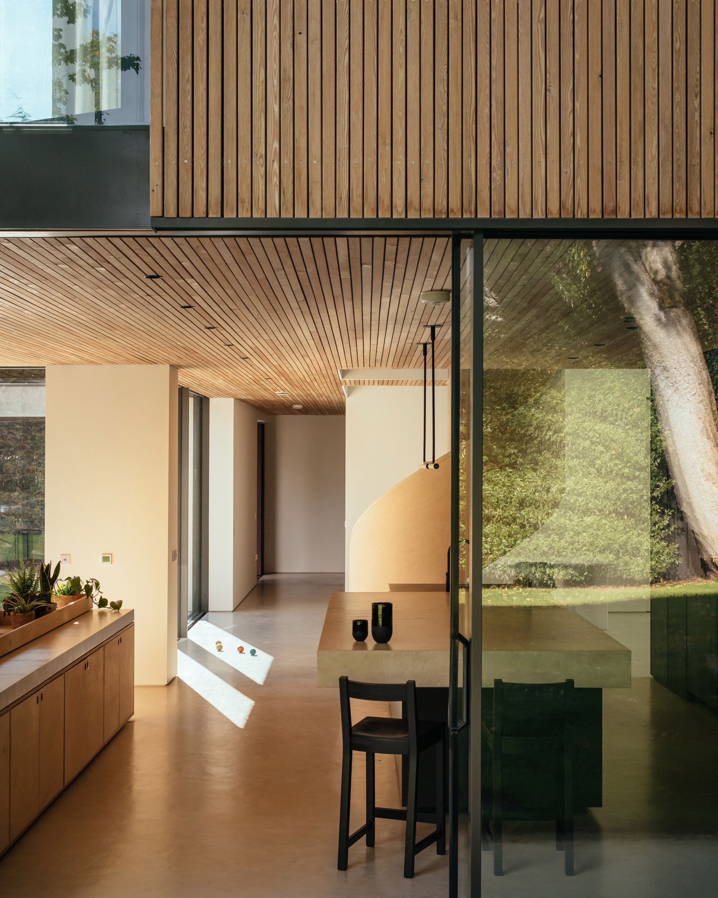 Peering from the outside into the kitchen area a modern house. A spiral birch plywood staircase can be seen in the background. Birch plywood cabinetry and a concrete island are in the foreground.