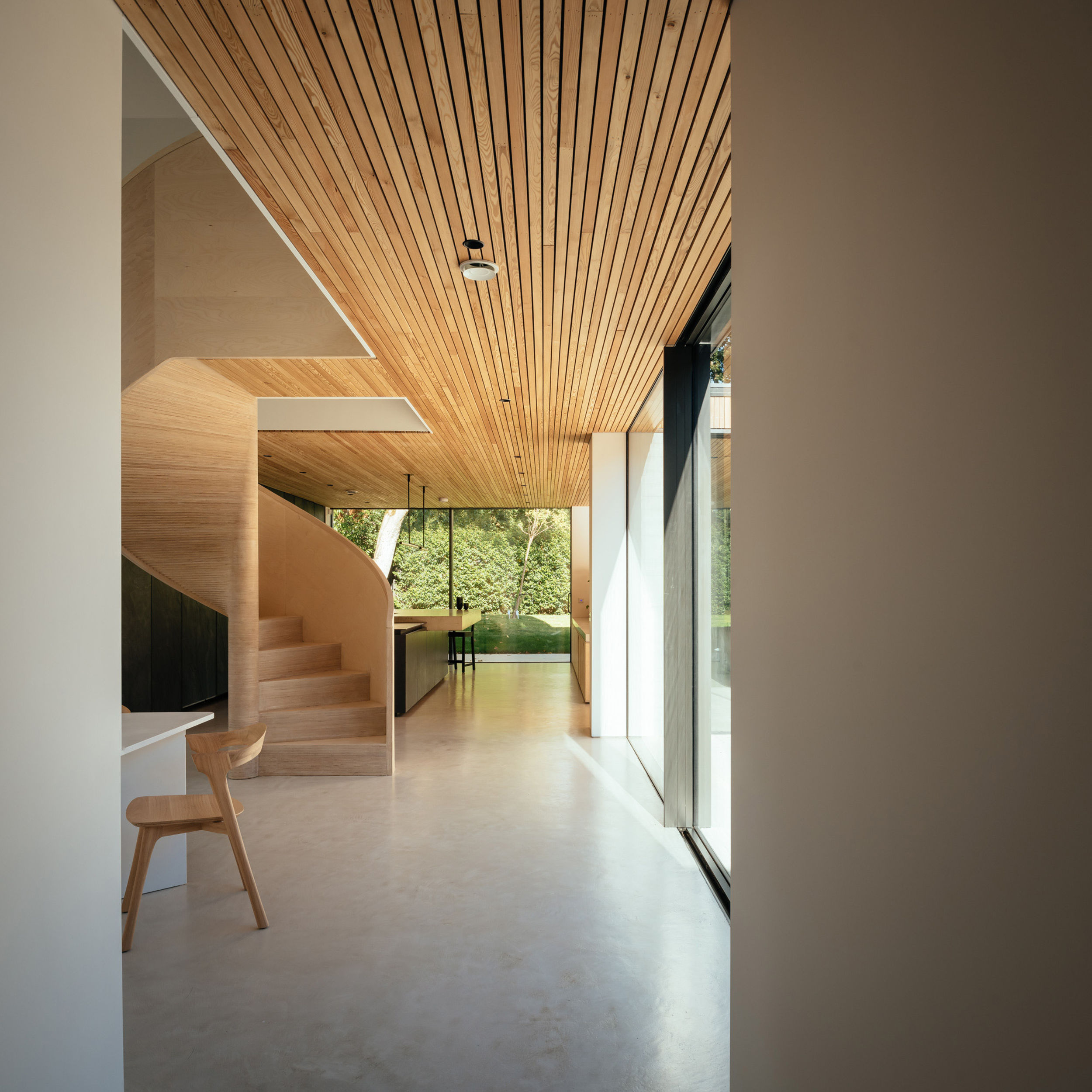 The entrance hallway of a modern house. The view takes us through to the dining and kitchen area. A birch plywood spiral staircase is in the centre of the space. Micro cement floors. Timber lined ceiling.