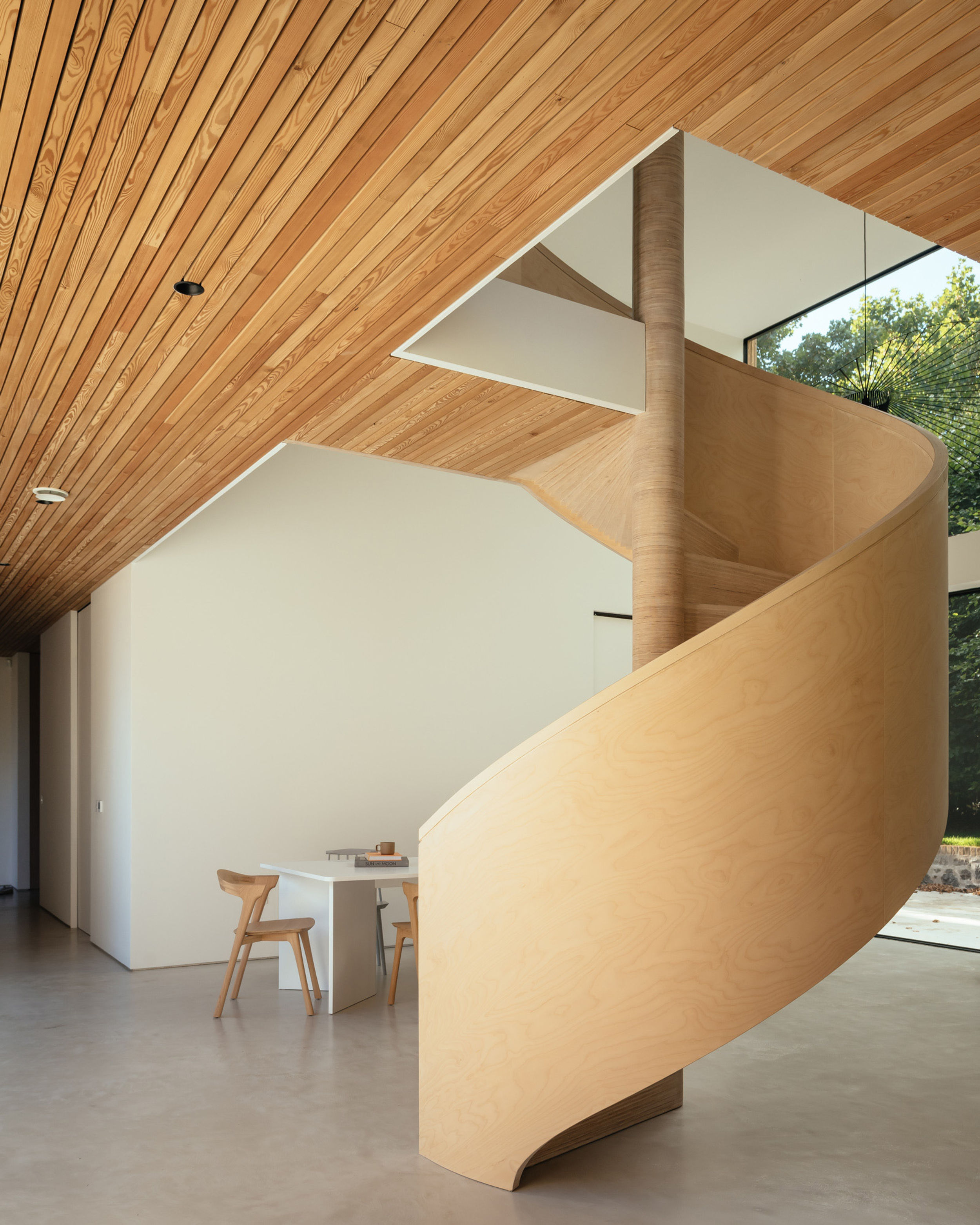 A spiral birch plywood staircase is the centrepiece of a room inside a modern house. Floor to ceiling glazing makes up the back wall. The floor is micro cement. The ceiling is timber lined. A white table and timber chairs sit in the background.