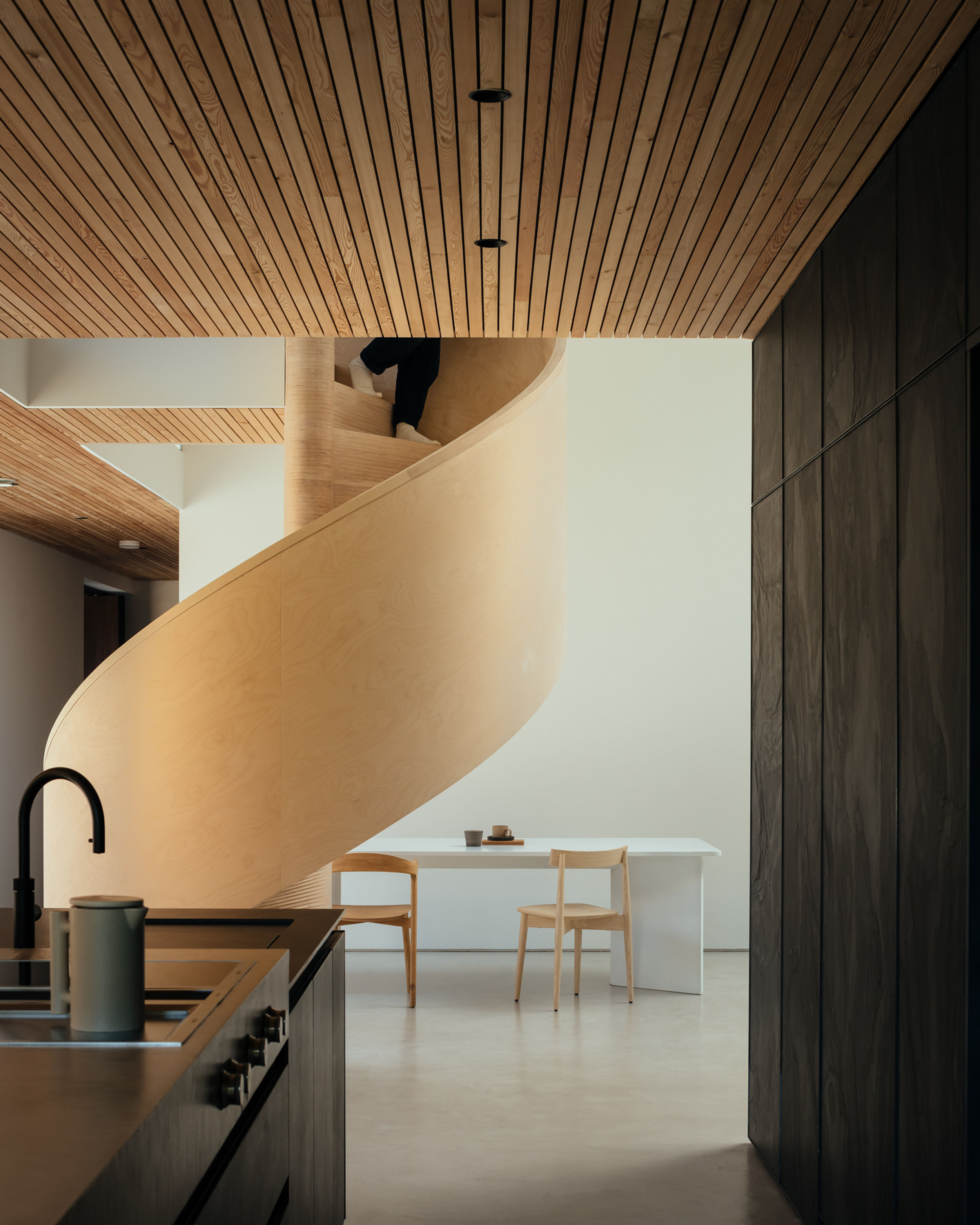 The view from the kitchen of a modern house. A black kitchen island with concrete worktop are in the foreground. The kitchen sink and taps can also be seen. In the background a persons feet can be seen descending a birch plywood spiral staircase. The floor is micro cement. A white table and timber chairs are also in the background.