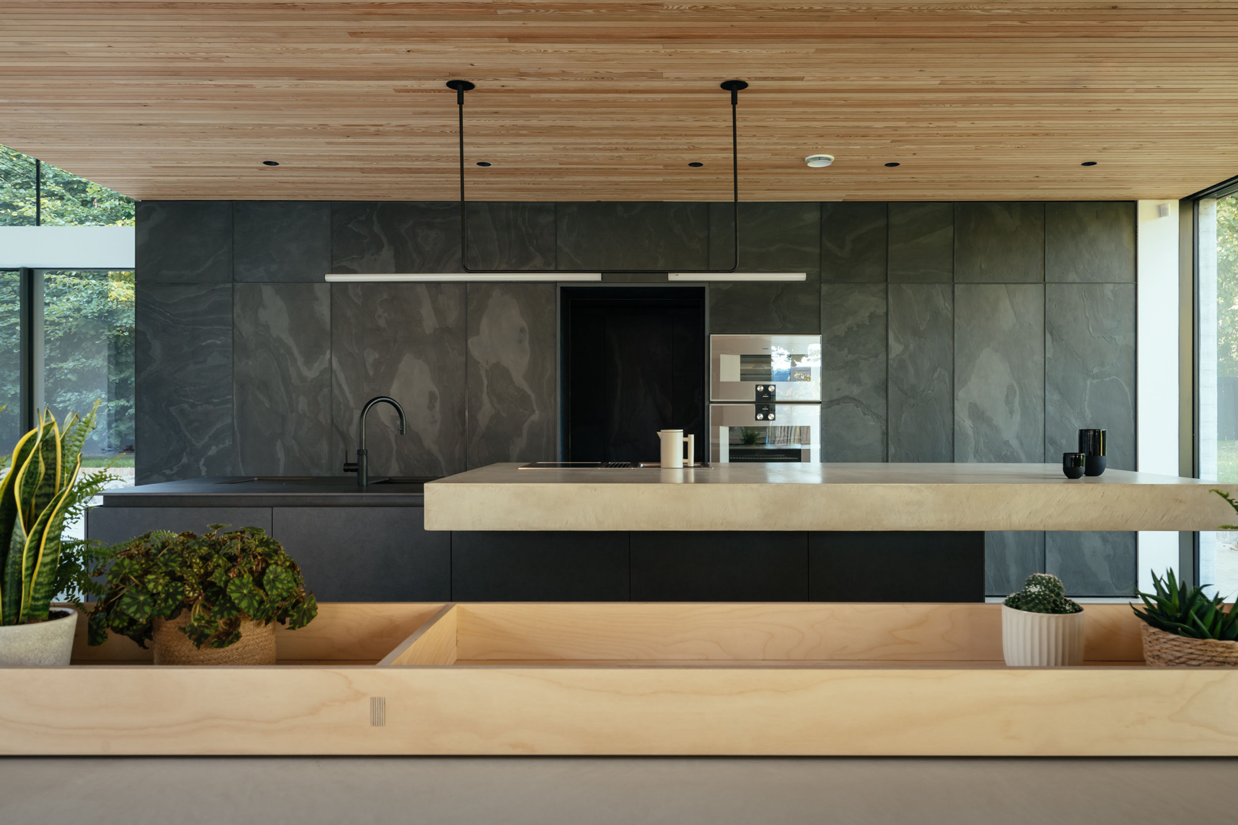 The kitchen and living area inside a modern house. Black kitchen cabinets line the back wall. A kitchen island sits in the centre of the room, with its concrete work top. Birch plywood cabinetry and a birch plywood spiral staircase can be seen. The floor is micro cement. The ceiling is timber lined.