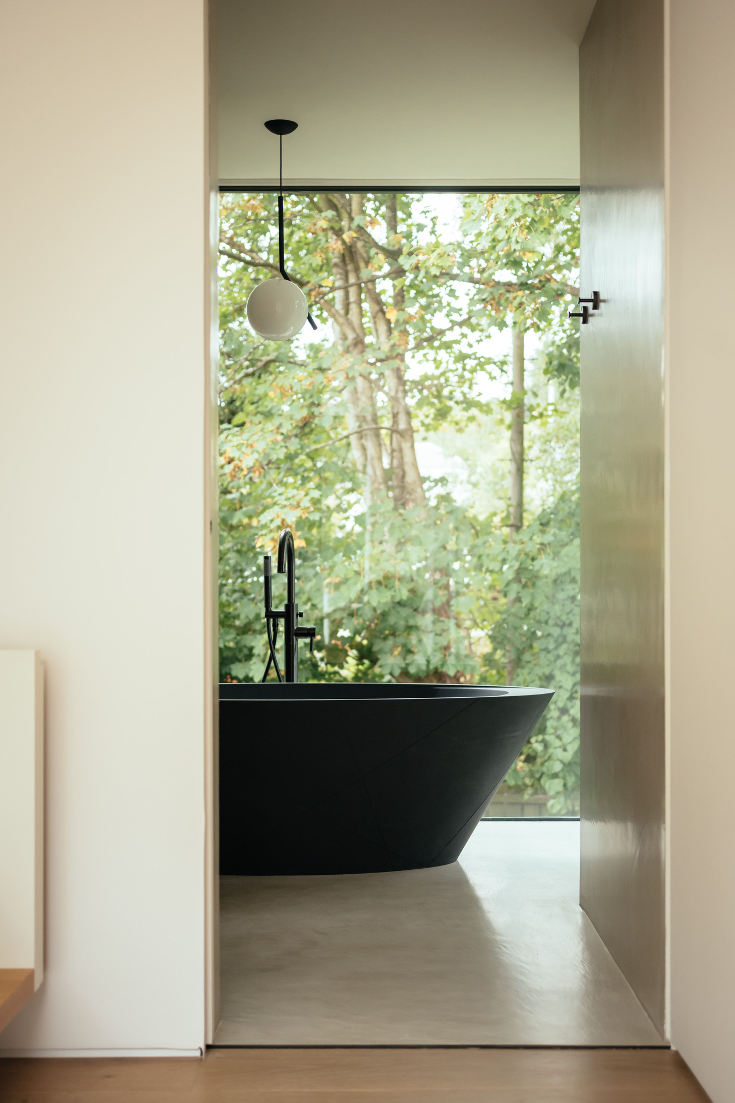 Looking into the ensuite bathroom of a modern house. A modern black bathtub sits in the centre of the room. Floor to ceiling glazing allows light to flood into the bathroom.