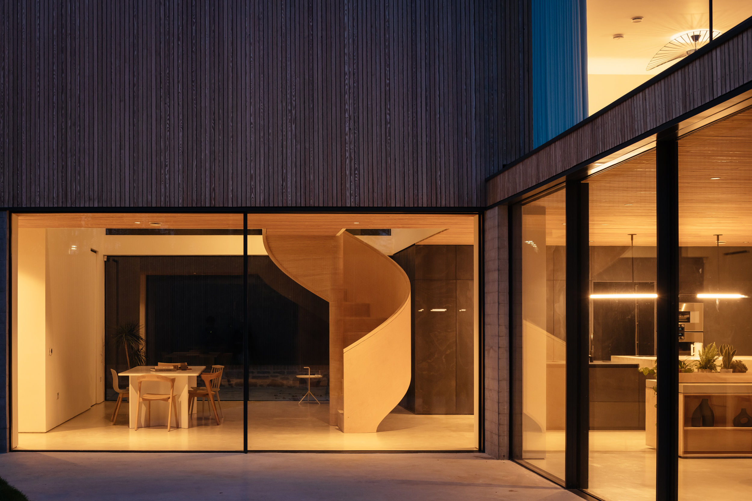 A twilight shot of a modern house, looking from the outside in. The lights are on inside the property and floor to ceiling glazing allows you to see right inside the home. A birch plywood spiral staircase is visible inside the home.