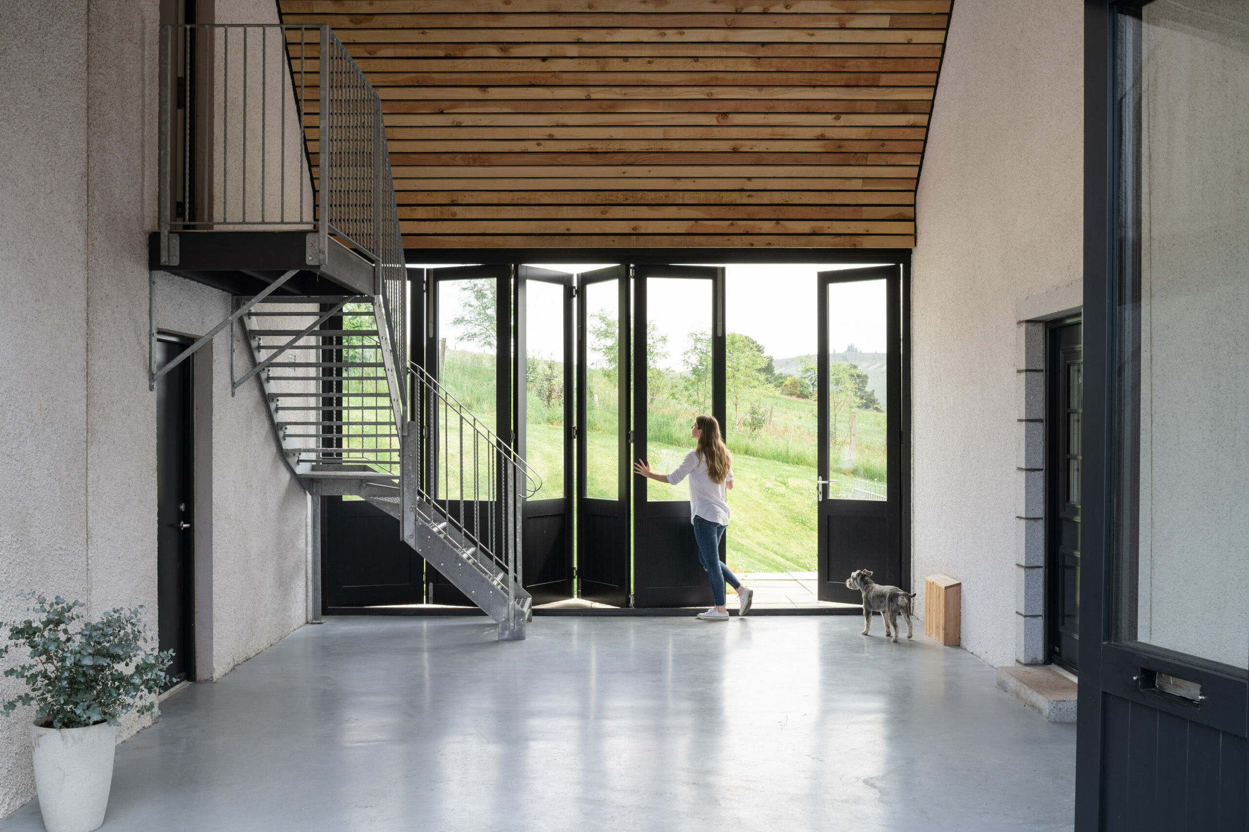 A woman pushing opening bifold doors in a carport. Timber linings on roof. Polished concrete floor.