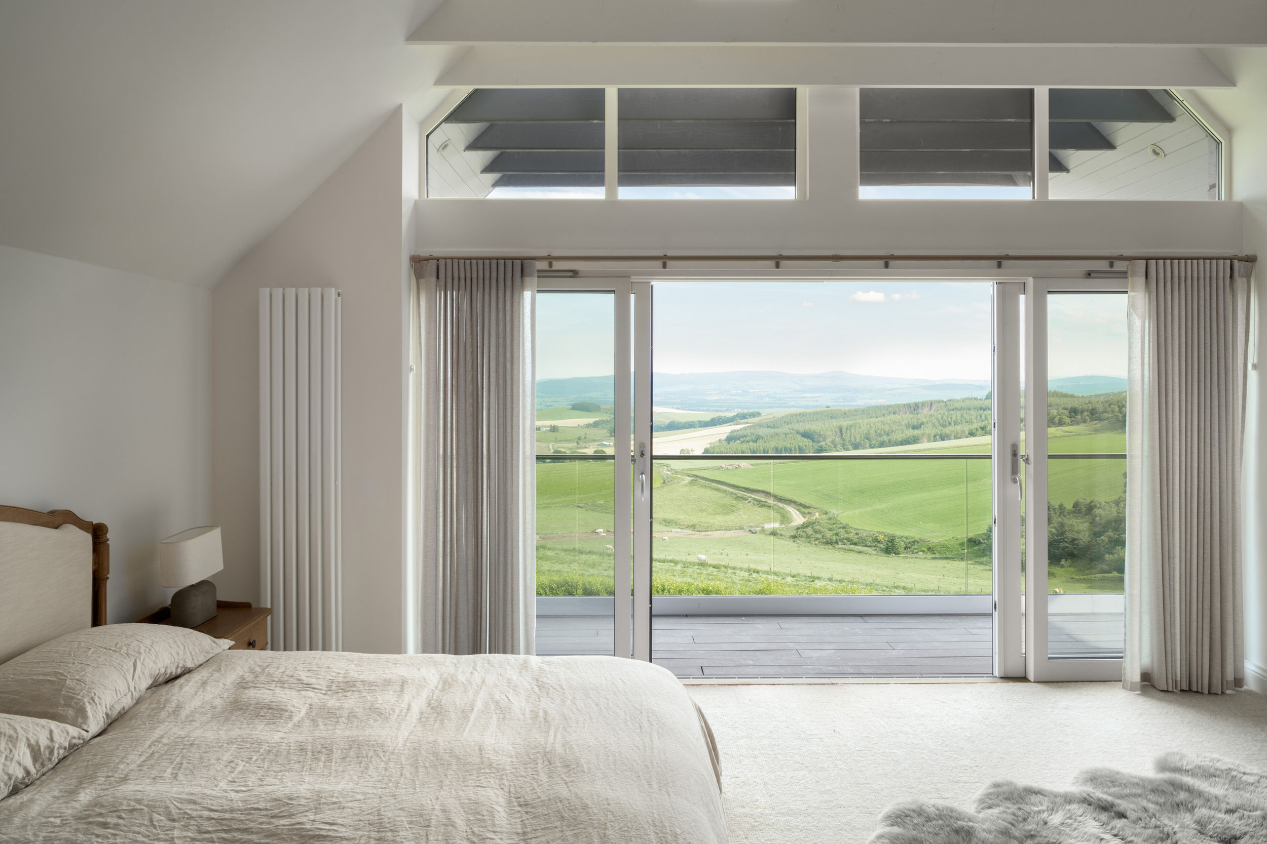 A master bedroom with a double bed with linen duvet. Exposed white ceiling ties. Floor to ceiling glazing shows off beautiful rural views. Balcony doors are open. Balcony has glass handrails and black timber flooring.