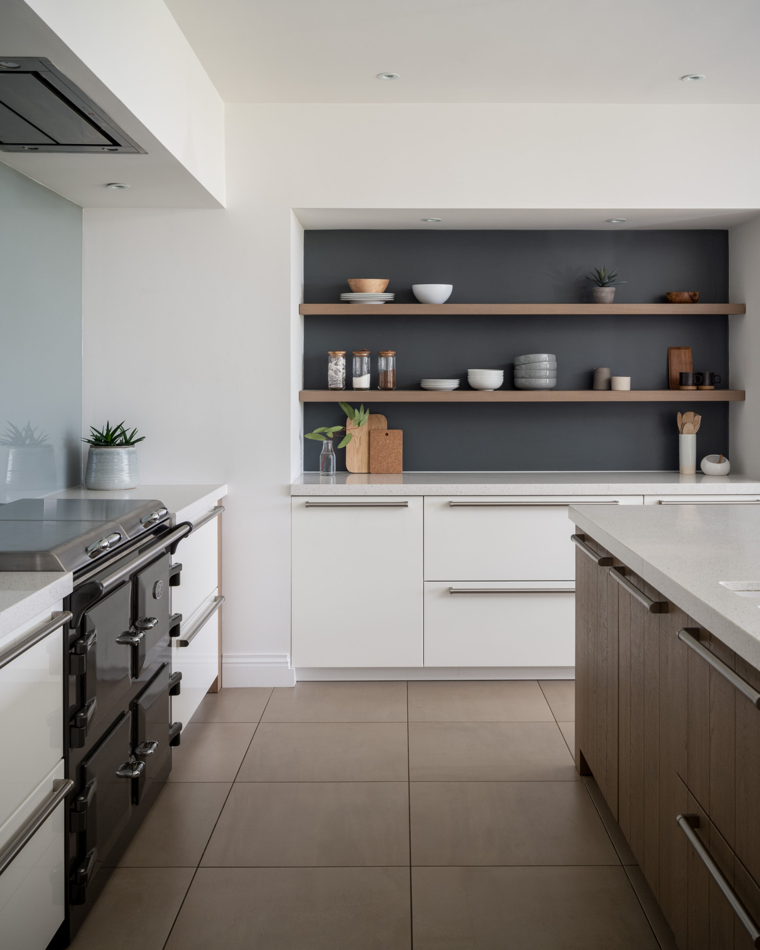 A kitchen scene. Bowls sit on two oak shelves, with a dark blue painted background. The kitchen counter tops are white marble. Large square beige tiles on the floor. Dark grey range cooker on the left of the room.