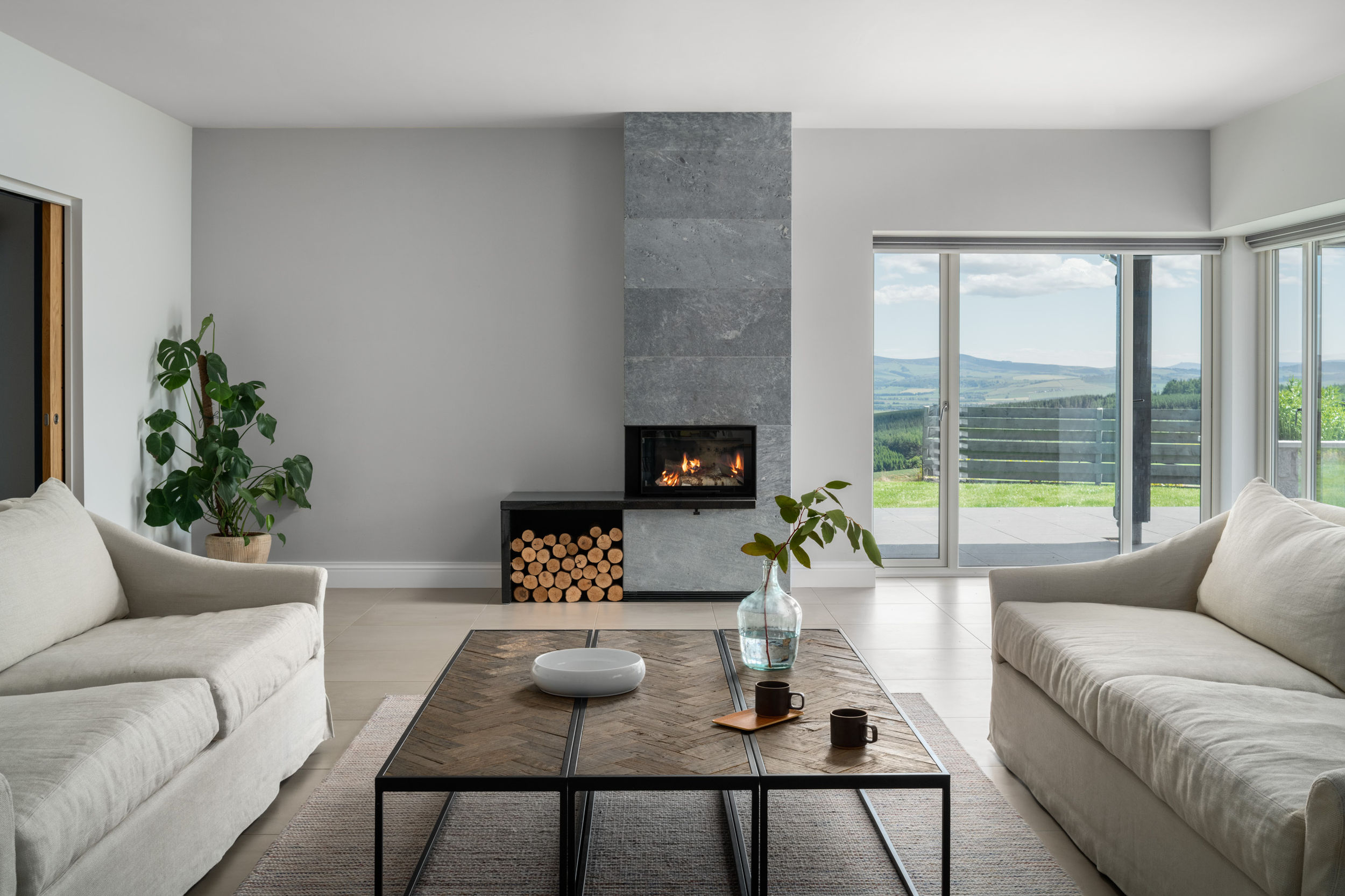 A living room interior with a grey stone fireplace on a back wall and two cream couches on each side of the room. A large wooden table sits on a wool rug in the centre of a room.