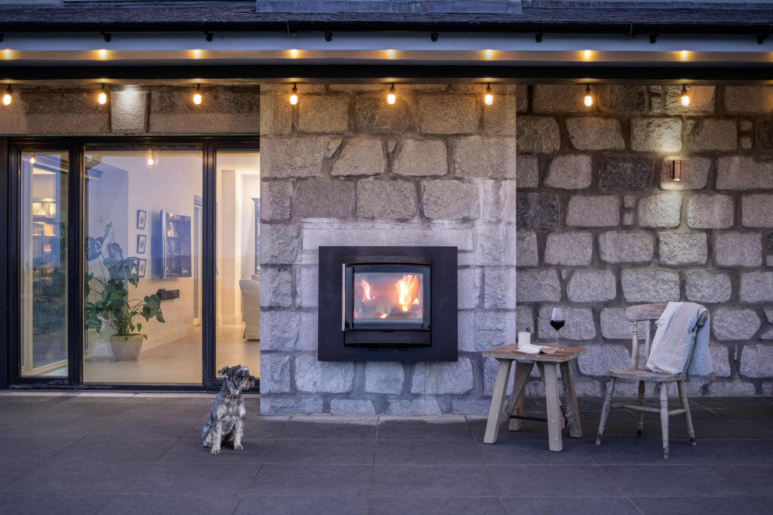 A built-in external fireplace burns outside a house as a dog watches on. The fireplace surround is grey granite stone. Party lights hang above the fireplace. A small wooden table and chair are next to the fire.
