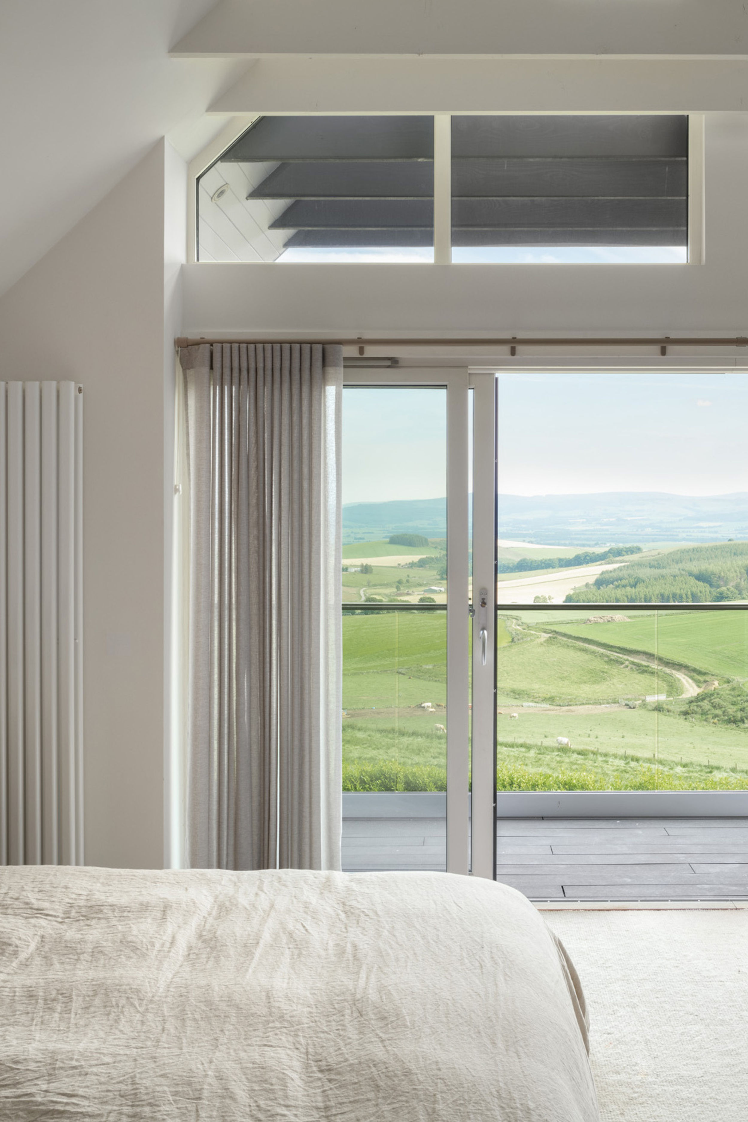 A master bedroom with a double bed with linen duvet. Exposed white ceiling ties. Floor to ceiling glazing shows off beautiful rural views. Balcony doors are open. Balcony has glass handrails and black timber flooring.