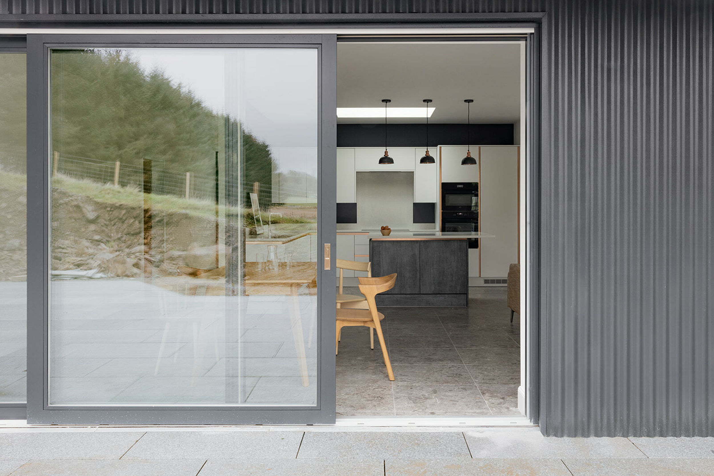 An exterior view of a house clad in grey corrugated sheeting. A sliding door is partially open, revealing a kitchen inside with a wooden dining table and chair.