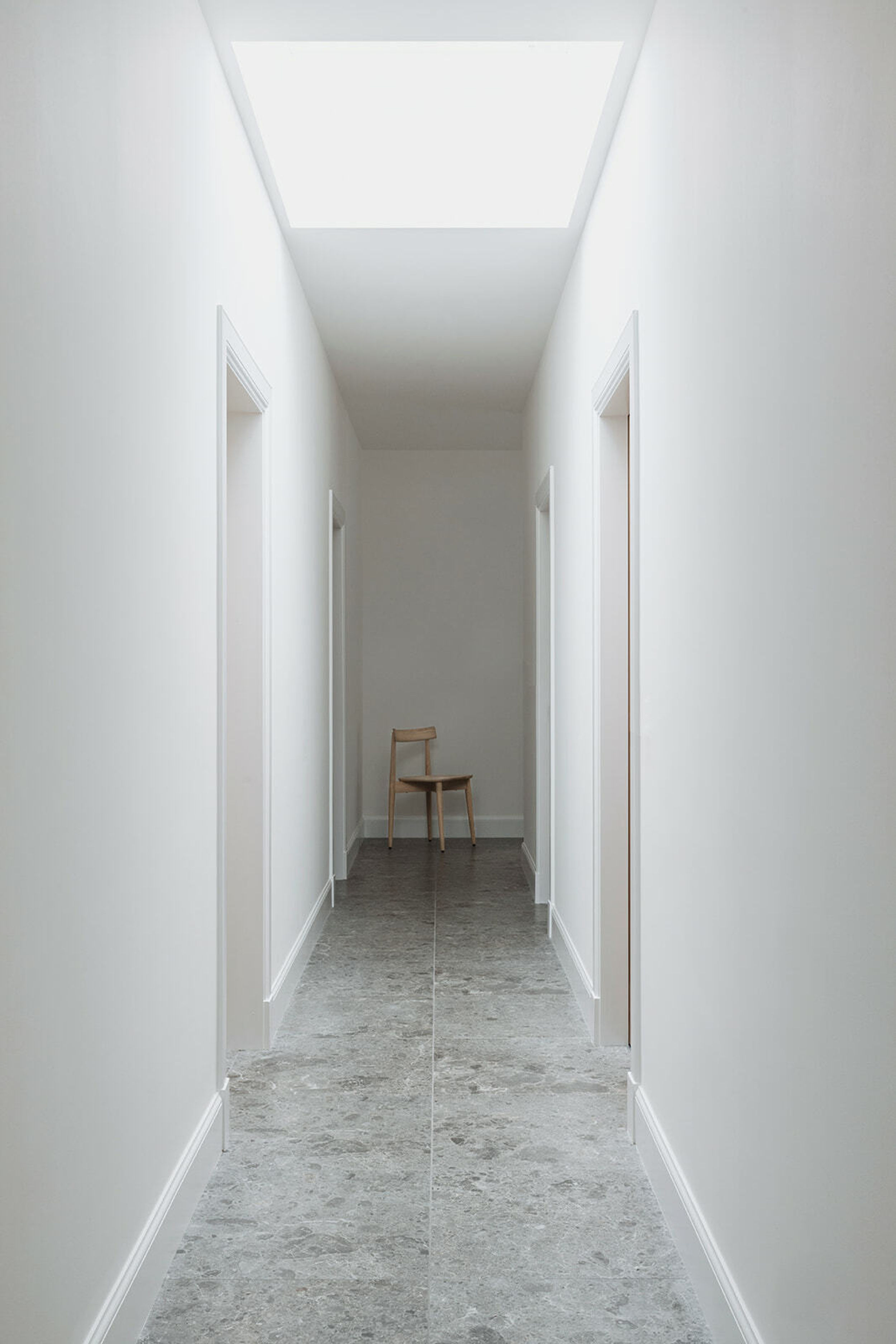 A long grey tiled corridor. White painted walls. Sunroof in the middle of the corridor shines light. A chair sits at the end of the corridor.