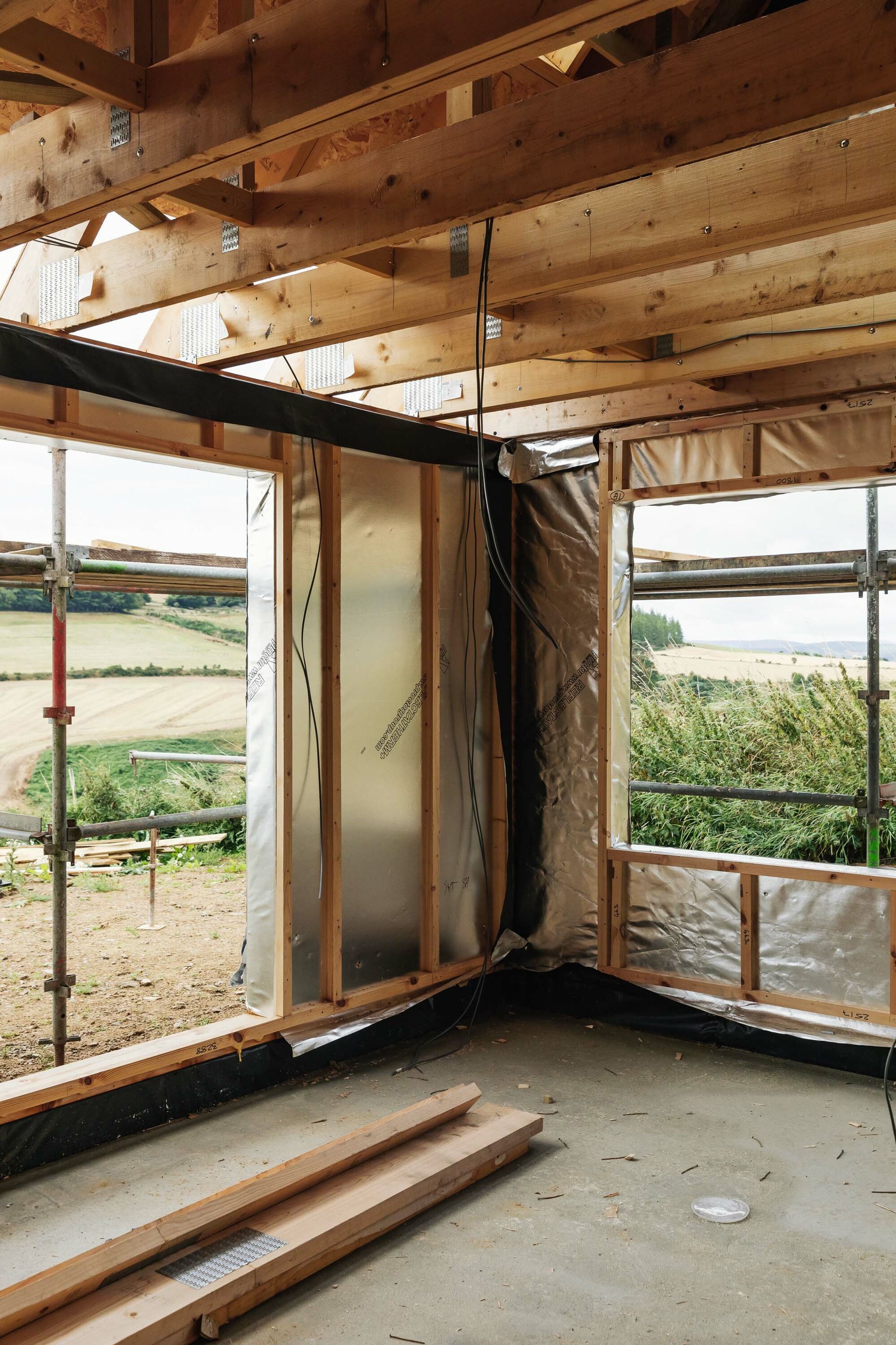 Inside a half-built timber frame home. Materials include Oriented Strand Board (OSB). One wall of the new house is covered in silver, windproof building paper.  Timber frames are exposed. Electrical cables hang from the ceiling. Sawdust covers the concrete floor. Barley fields are in the distance.